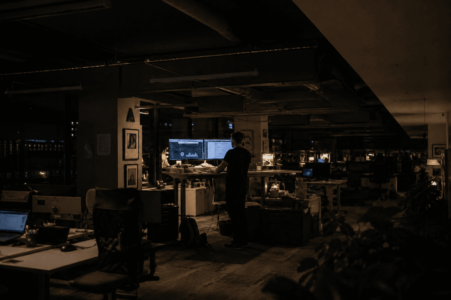 A wide view of a dimly lit open-plan office at night, with a lone engineer small within the frame working at a standing desk surrounded by rows of glowing monitors.