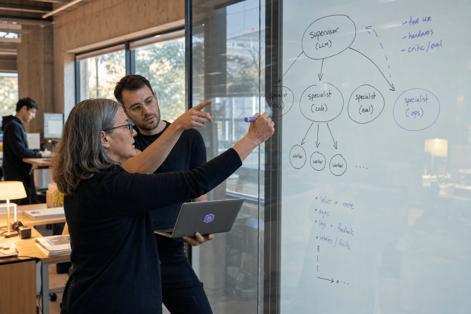 A senior female ML engineer draws a hierarchical node diagram on a glass whiteboard while a male colleague with an open laptop points at the sketch, in a daylit Australian tech office with exposed concrete walls and standing desks in the background.