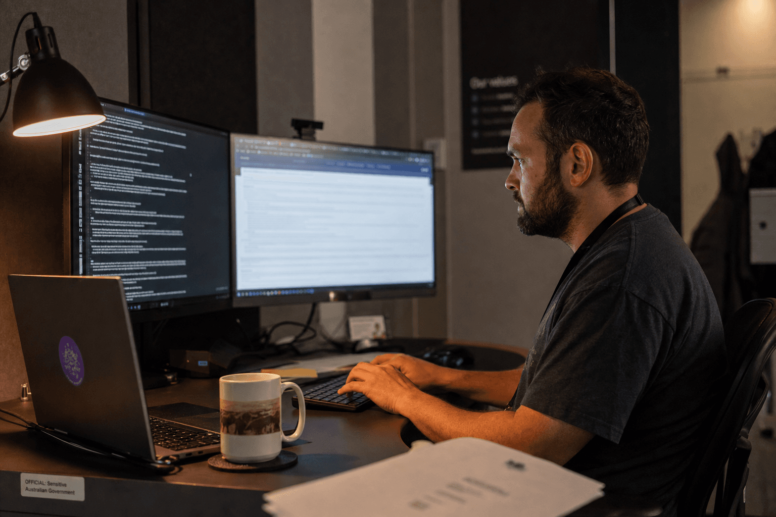 A male data engineer sits at a government office workstation at night, his face lit by the glow of dual monitors and a warm desk lamp, surrounded by printed documents and policy papers in a dim Australian tech workspace.