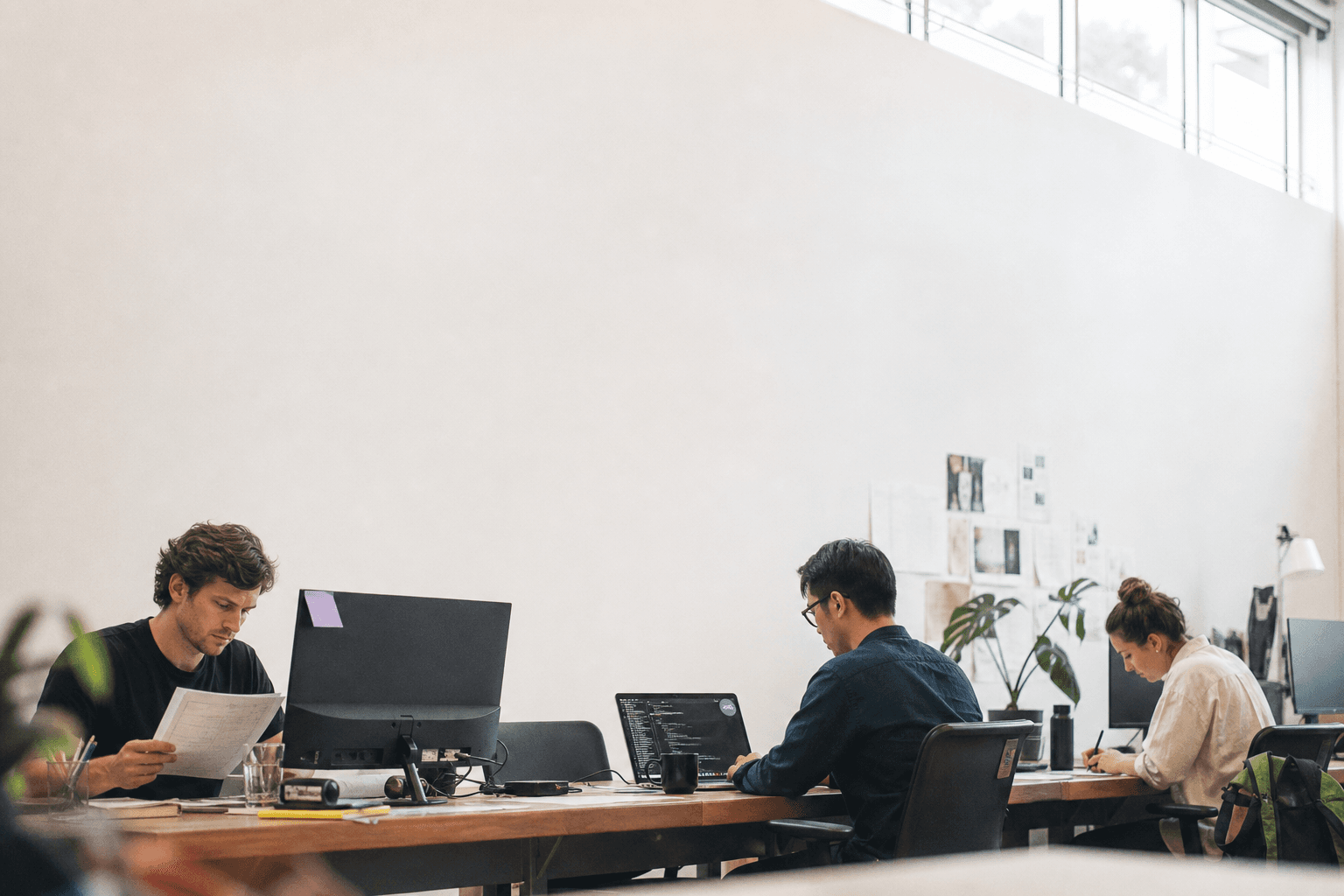 Three developers sit spaced along a long shared desk in a bright Australian tech office, each working differently — reviewing a contract, using a laptop terminal, and sketching on a notepad — beneath a wide expanse of pale daylit wall.
