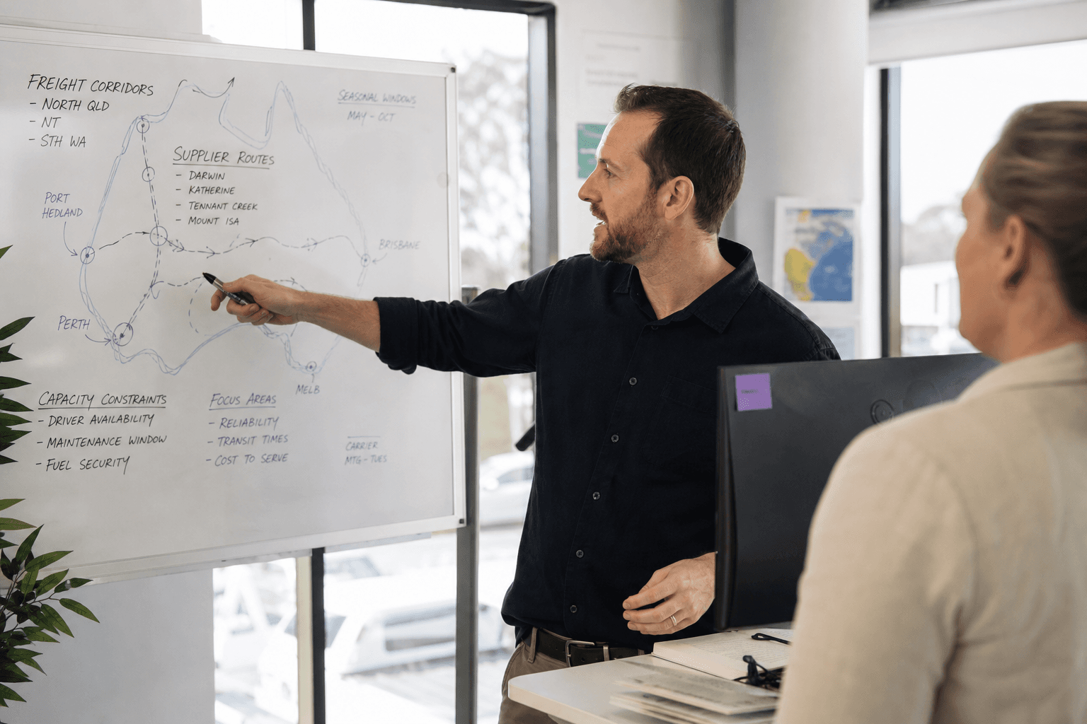 Side profile of a logistics planner standing at a desk, gesturing toward a whiteboard with route sketches, in a bright office with natural daylight.
