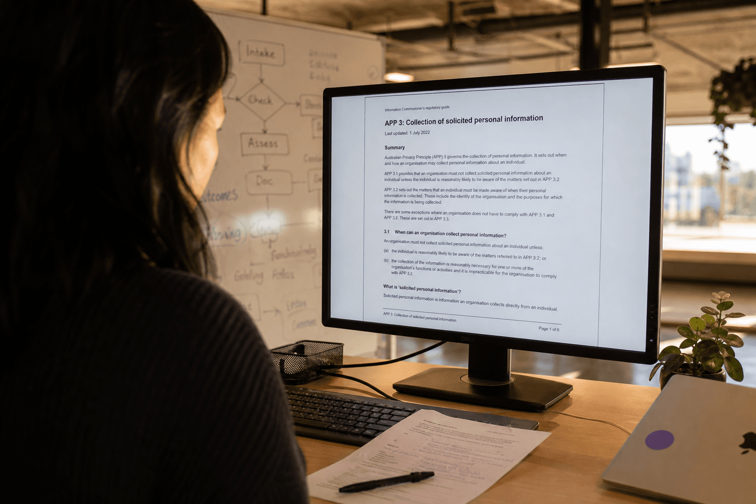 Over-the-shoulder view past a woman at a standing desk reviewing a regulatory document on a large monitor, warm golden-hour light from an office window mixing with screen glow, handwritten notes visible on a printed page beside the keyboard, whiteboard with flow diagrams blurred in the background.