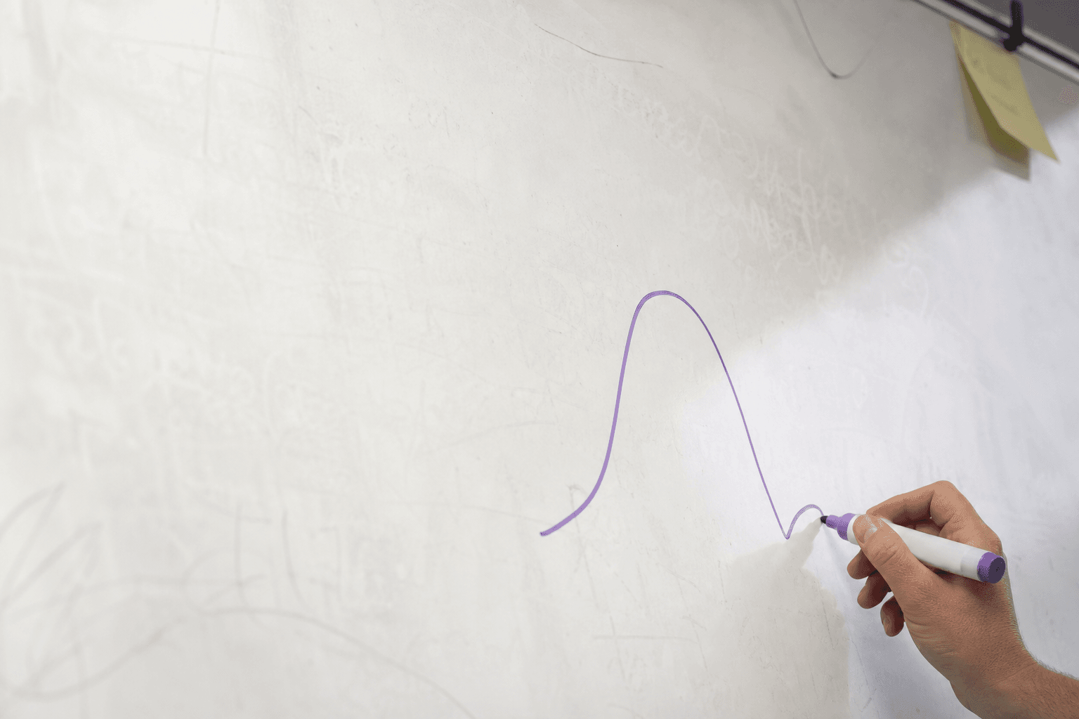 Extreme close-up of an off-white whiteboard in an Australian office, a single hand drawing a small looping curve in violet marker, the pale surface filling most of the frame with a faint sticky note visible at the edge.