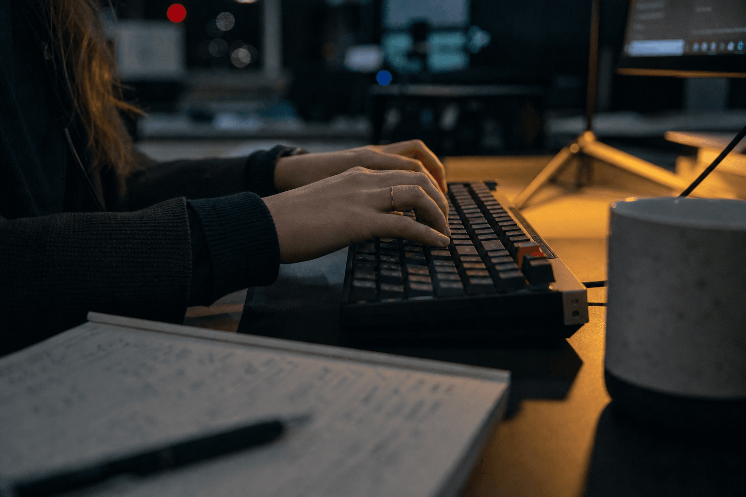 Close-up of a woman's hands typing on a mechanical keyboard in a dimly lit office, lit primarily by cool screen glow and a warm task lamp, with a blurred bokeh of monitor light in the background.