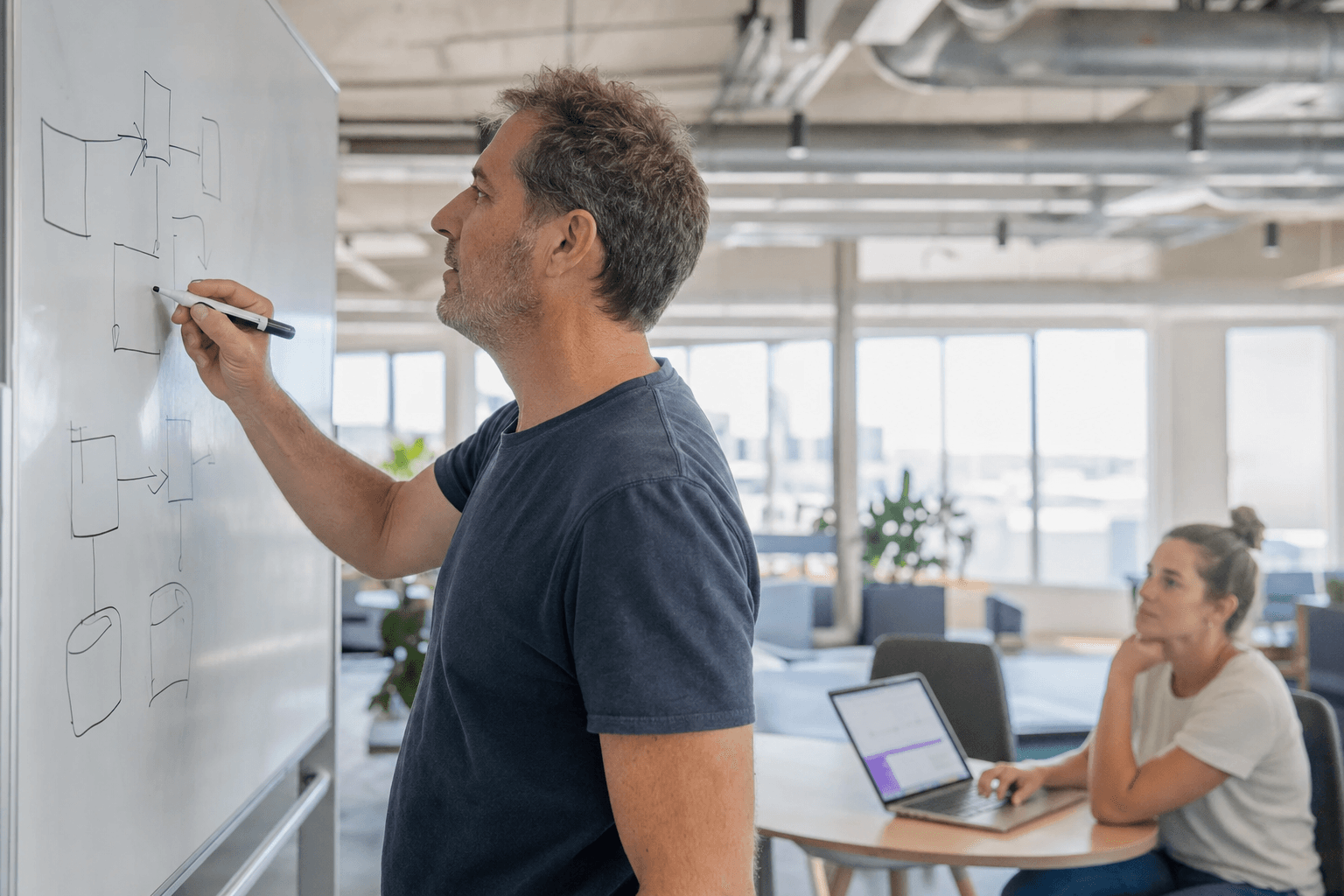 A man in his fifties stands in side profile at a whiteboard sketching a process diagram with a marker in a bright daylit Australian office, while a younger woman seated at a table in the background looks on with a laptop open.