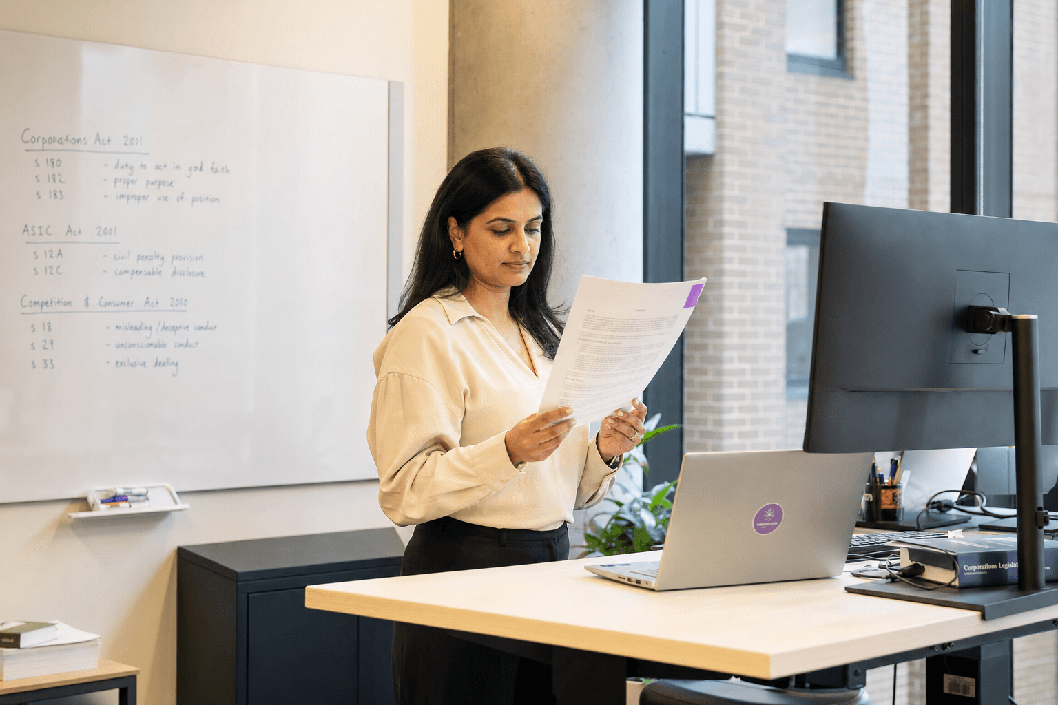 A woman reads a printed legal document at a standing desk in a light-filled Australian office, with a whiteboard of handwritten notes and a daylit window receding in depth behind her.
