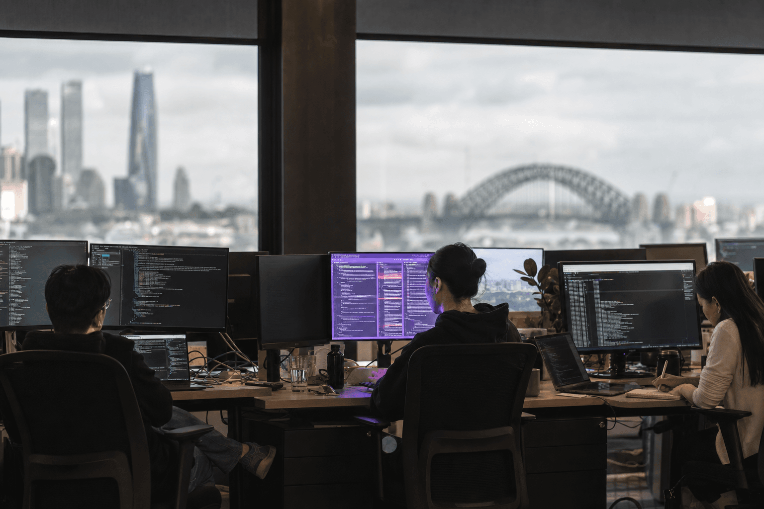Three developers sit in silhouette along a wide workstation bench in a daylit Australian office, with a bank of monitors showing code and terminal output, and a single violet-tinted screen casting a glow on the central engineer's face against a softly defocused city window.