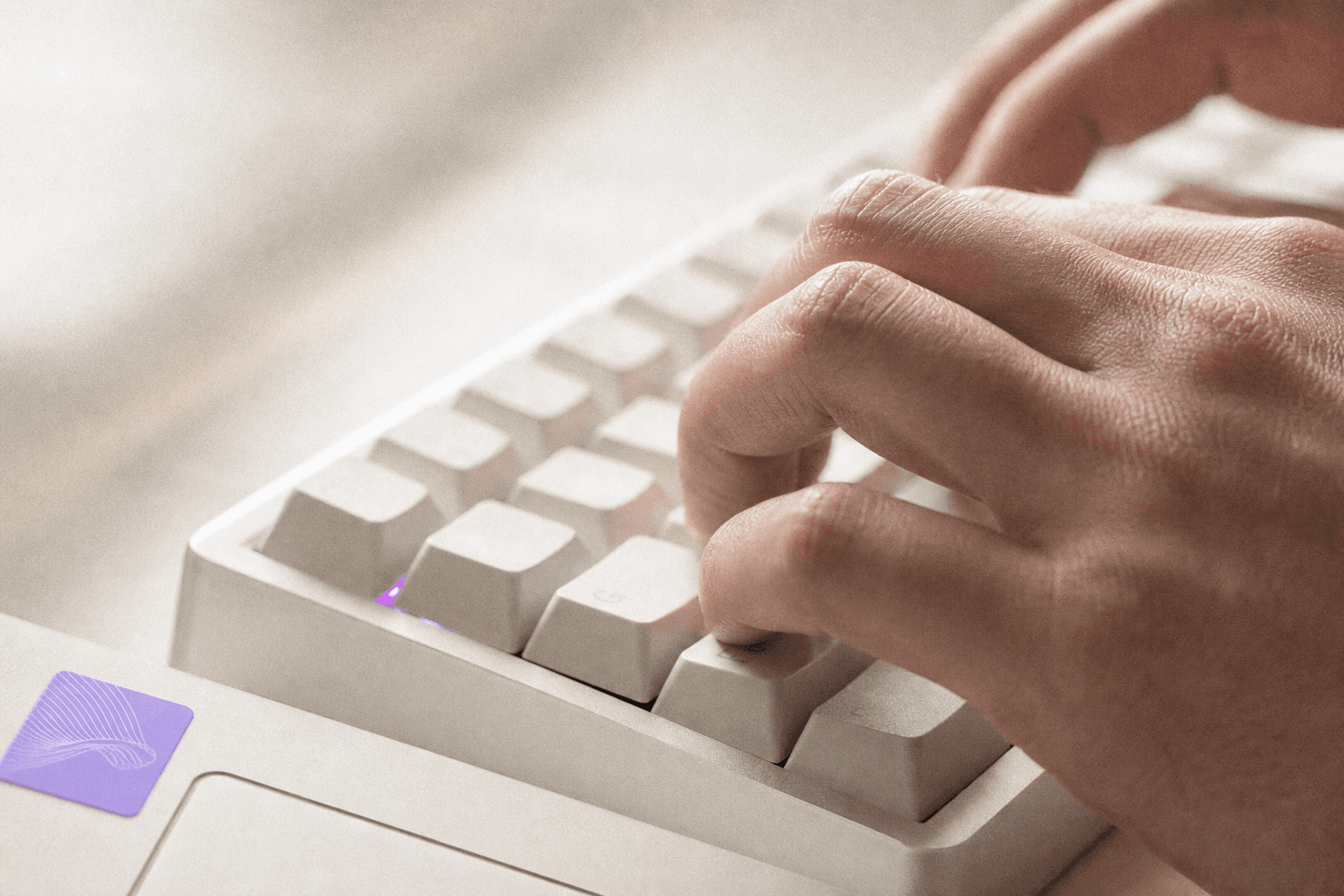 Extreme close-up of a developer's hands on a mechanical keyboard, pale desk surface filling most of the frame, a small violet laptop sticker providing the only colour accent, raking natural window light highlighting keycap texture.