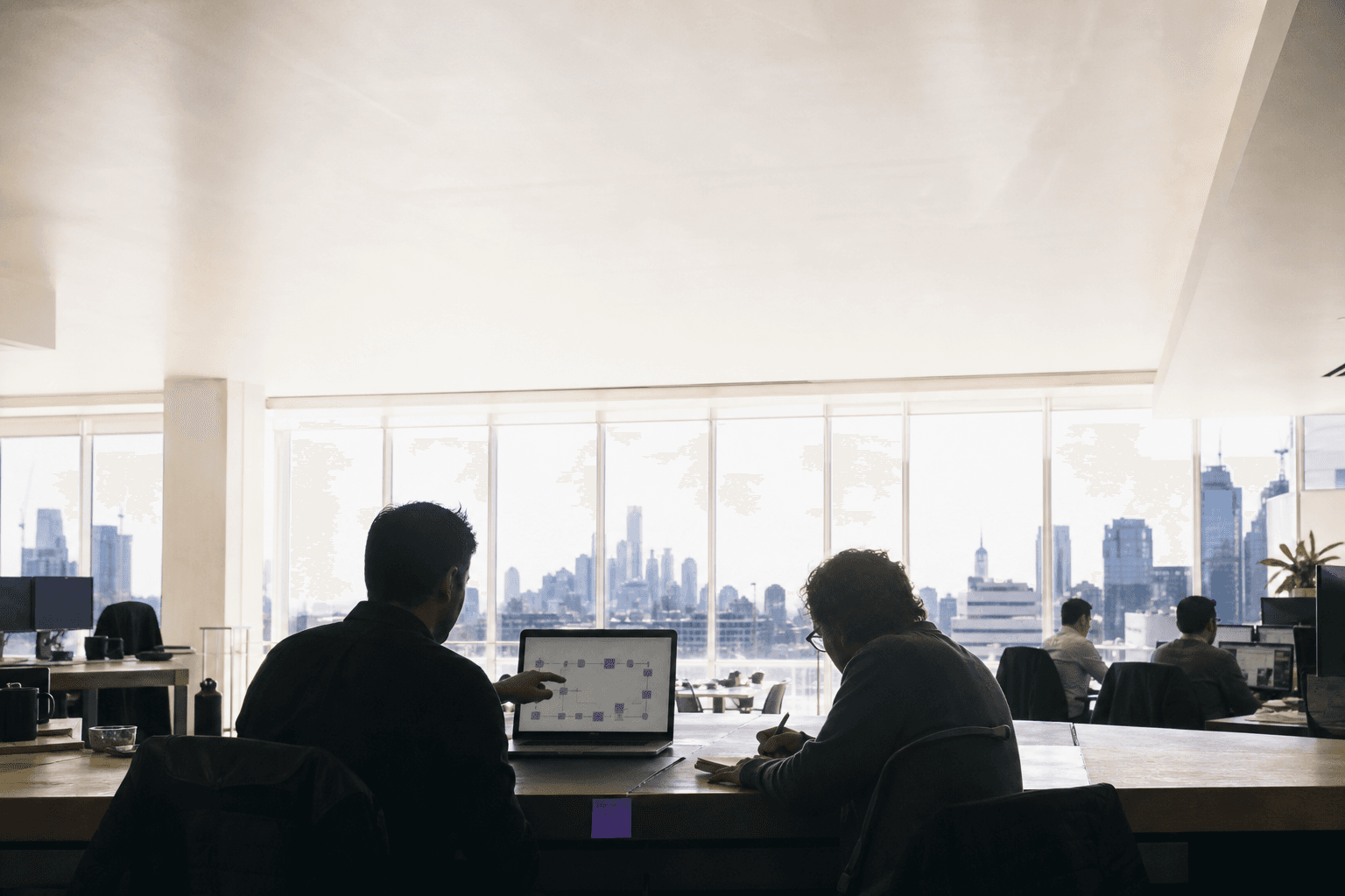 Two software architects sit in silhouette at a long bench in a bright Melbourne office, backlit by large windows flooding the frame with white daylight, one pointing at a laptop showing a schematic diagram while the other sketches on paper.