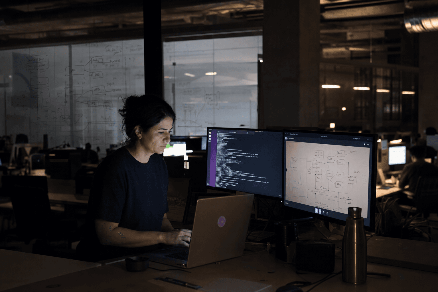A woman at a standing desk in a dark Australian open-plan tech office, multiple monitors glowing, with layered planes of glass partitions covered in whiteboard sketches and warm desk lamps receding into the background.