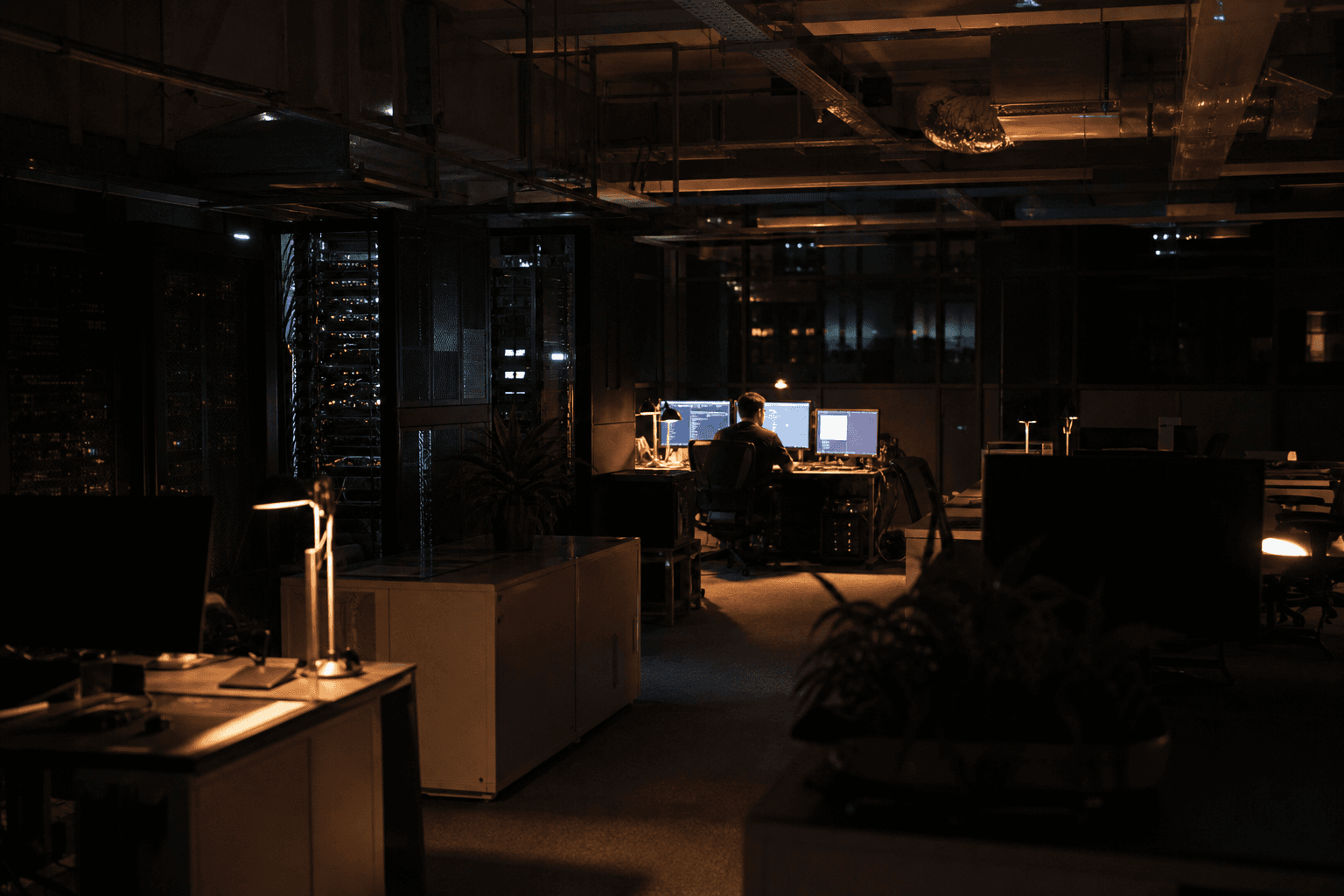 Wide view of a dimly lit Australian office at night with rows of desks creating separate zones, a lone engineer working at a glowing multi-monitor workstation in the distance.