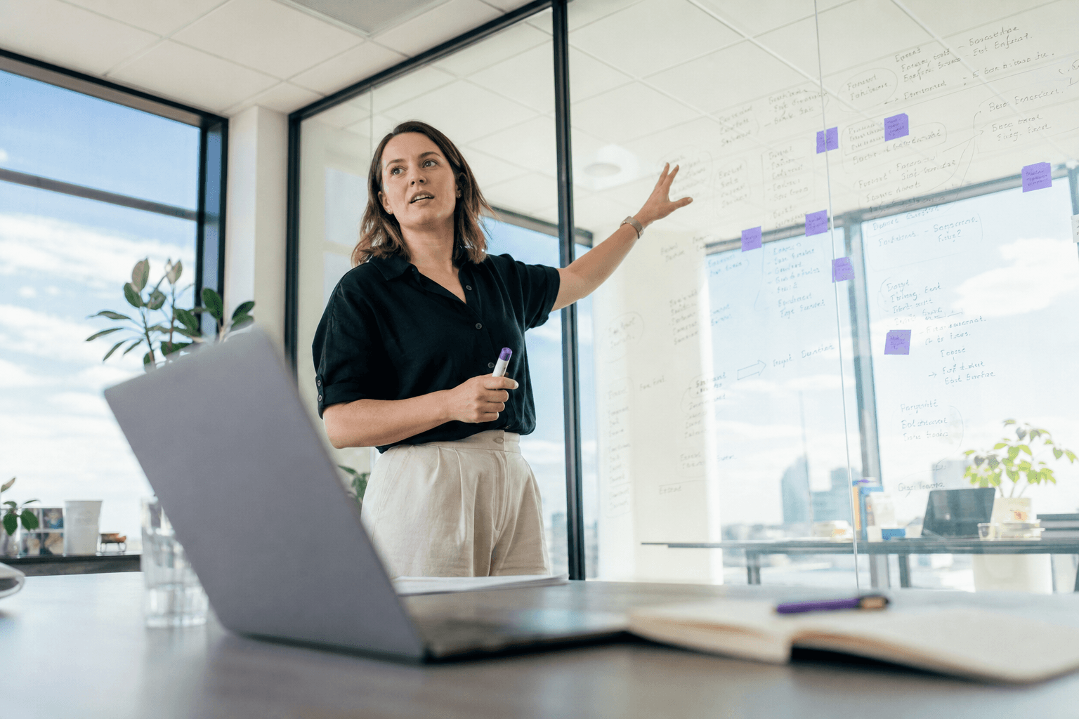 A person stands and gestures toward a glass wall covered in sketched notes and sticky notes in a bright, sunlit office, seen from a low angle past a laptop on a table.