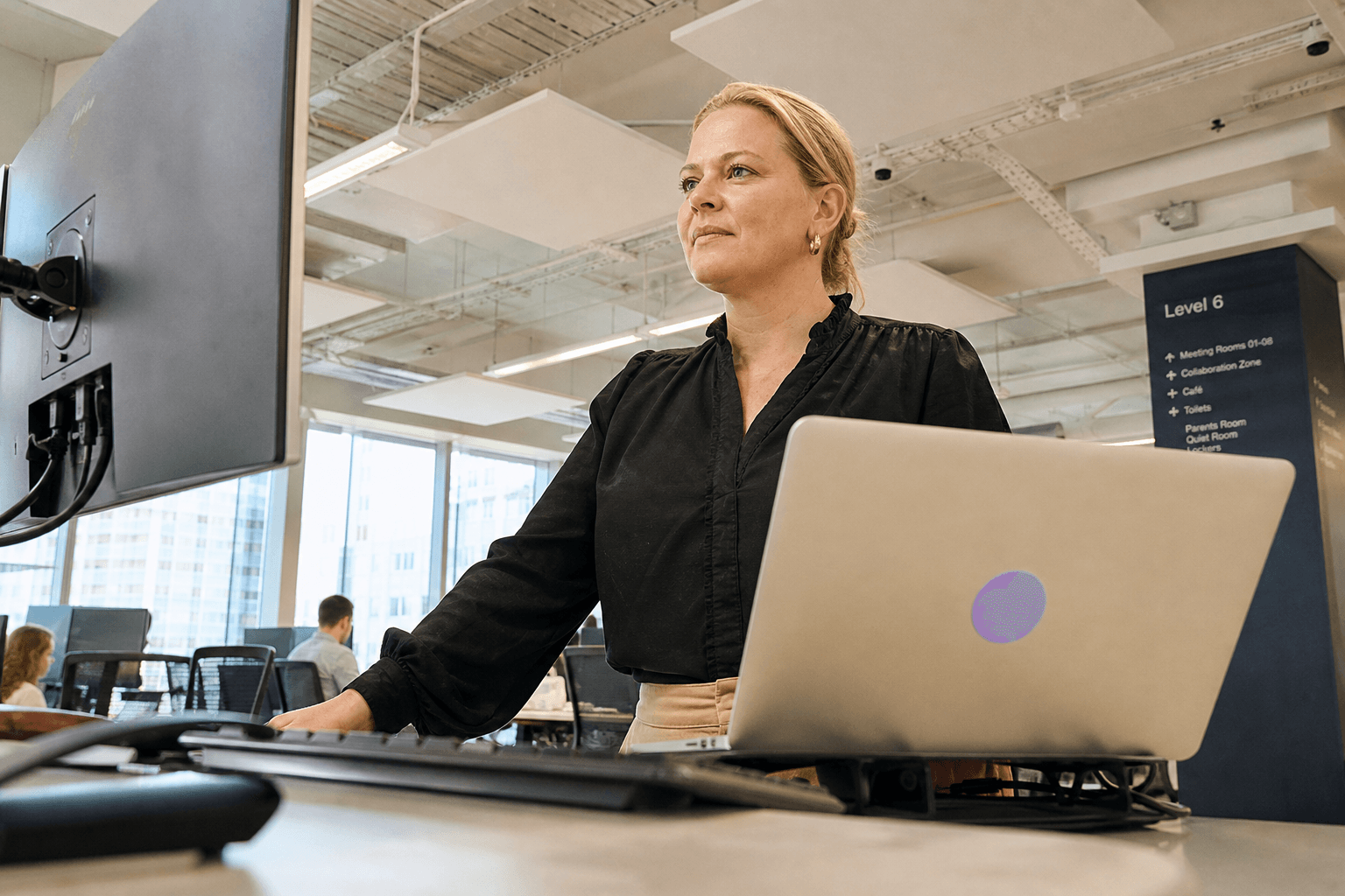 Low-angle photo looking up past a keyboard toward a fraud analyst standing at a desk reviewing an account activity screen, bathed in bright daylight in an open-plan office.