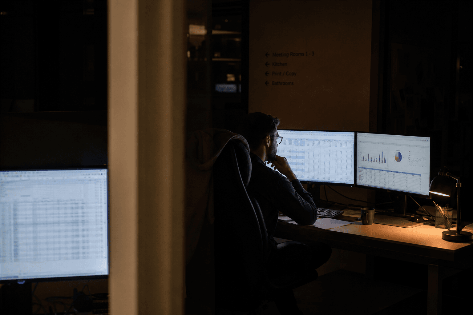 A data engineer working late at dual monitors validating reporting data, viewed through an office doorway, lit by screen glow and a warm desk lamp in a dim room.