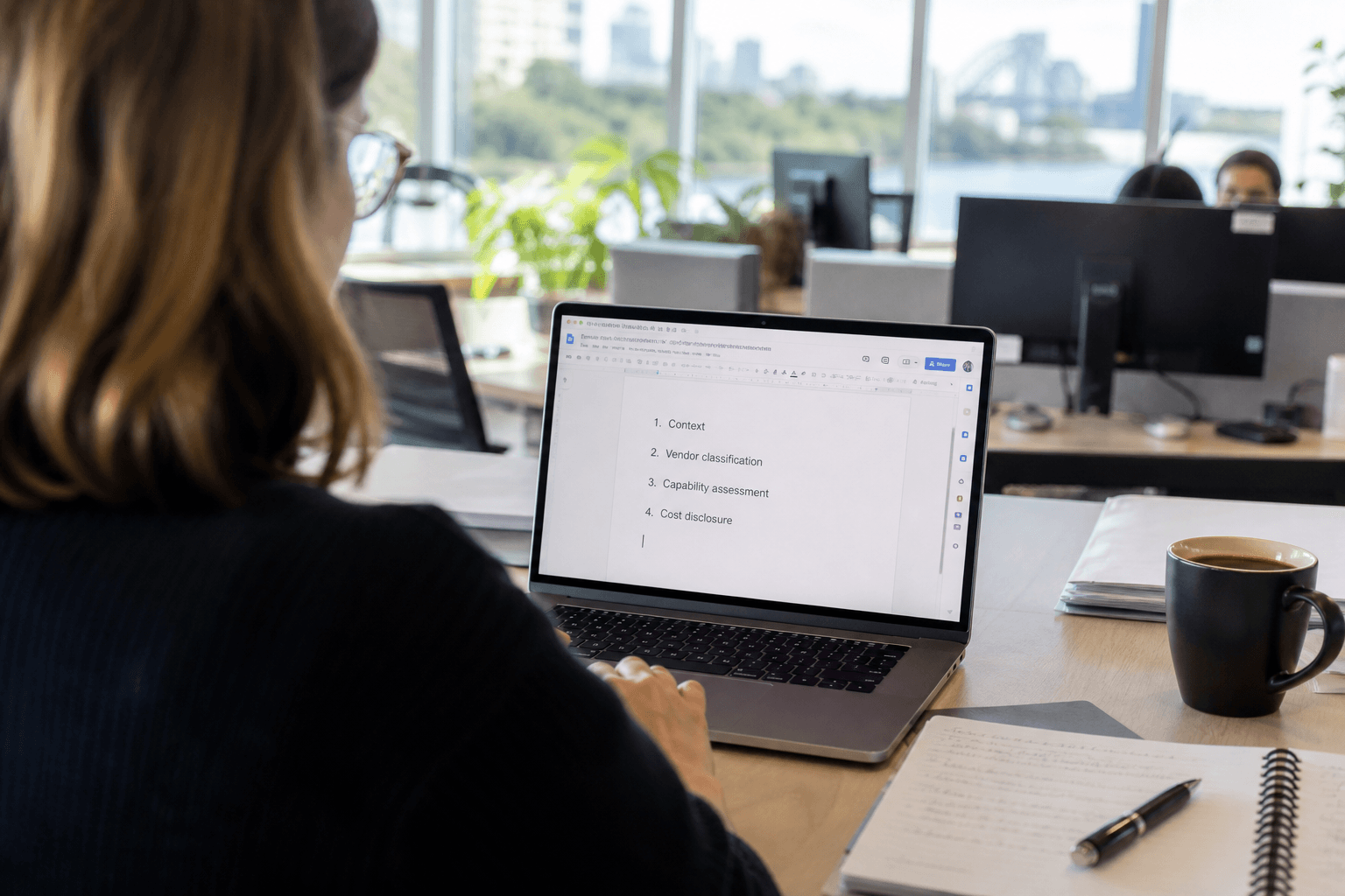 Over-the-shoulder view of someone typing a structured document outline on a laptop at a bright, daylit office desk with a notebook and coffee cup nearby.