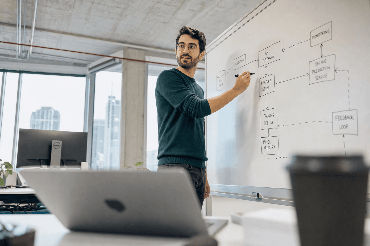 Low-angle view from a desk surface looking up past a laptop in the foreground to a male engineer standing at a whiteboard drawing a system architecture diagram, with bright natural daylight from large office windows in the background.