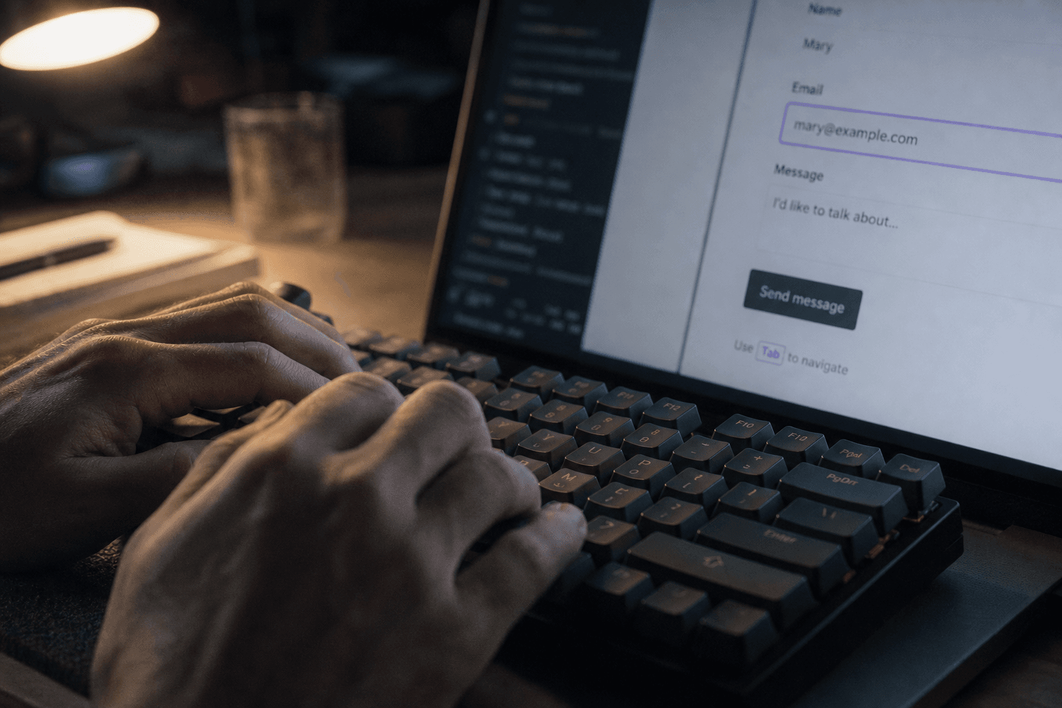 Close-up of hands typing on a keyboard while navigating a web form using the Tab key, with a laptop screen glowing in a dimly lit room.