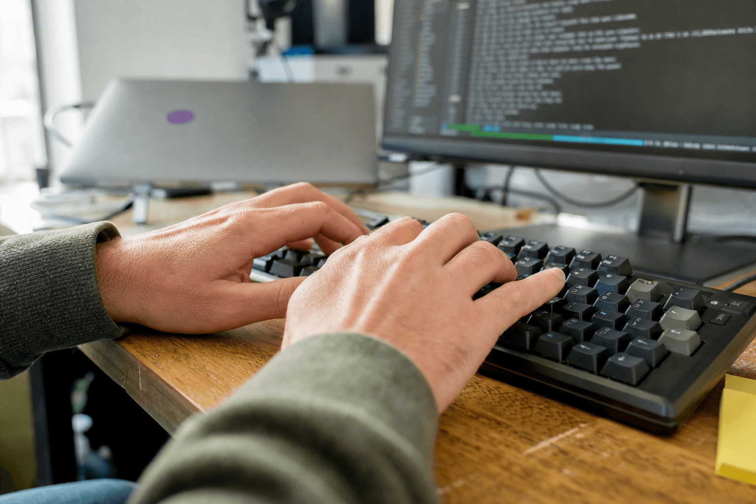 Close-up of a software engineer's hands typing on a mechanical keyboard at a timber desk, bright natural daylight from a window illuminating the scene, with a blurred terminal window glowing softly on a monitor in the background.