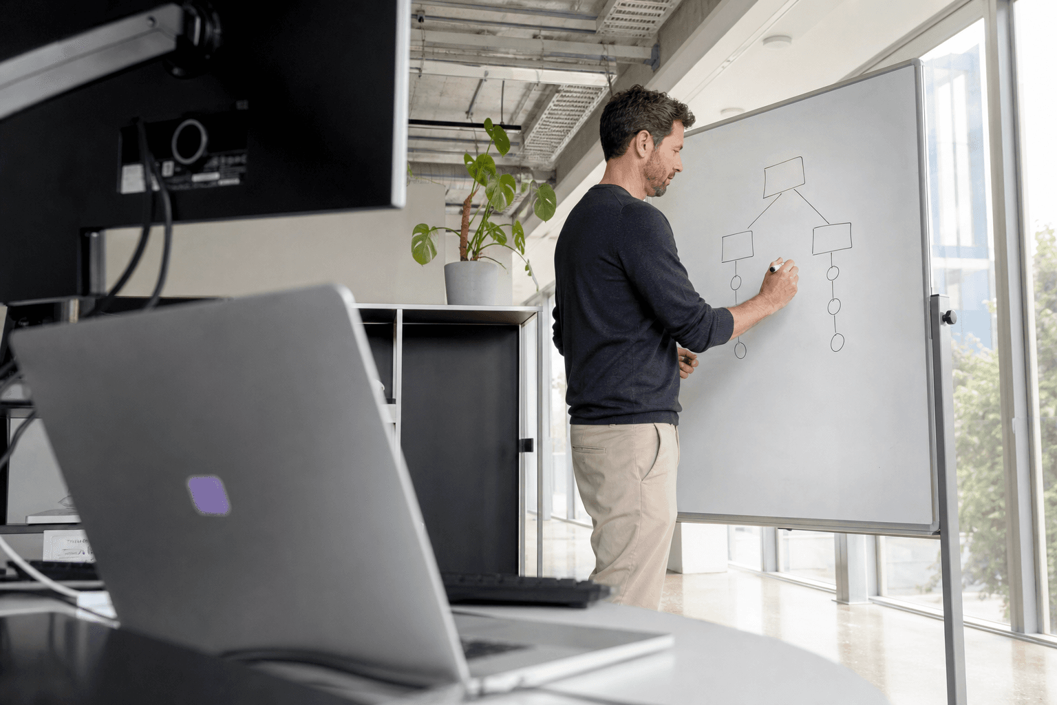 A person stands at a whiteboard sketching a simple decision diagram, viewed from a low angle past desk equipment, in a bright naturally lit office.
