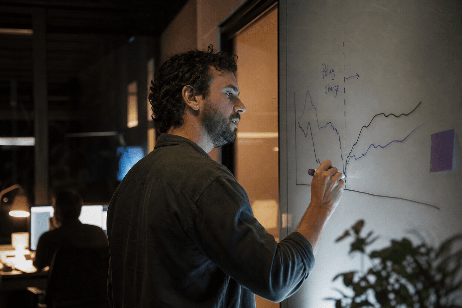 A man in profile sketches a before-and-after line chart on a glass whiteboard in a dimly lit office, illuminated by screen glow and a warm desk lamp.