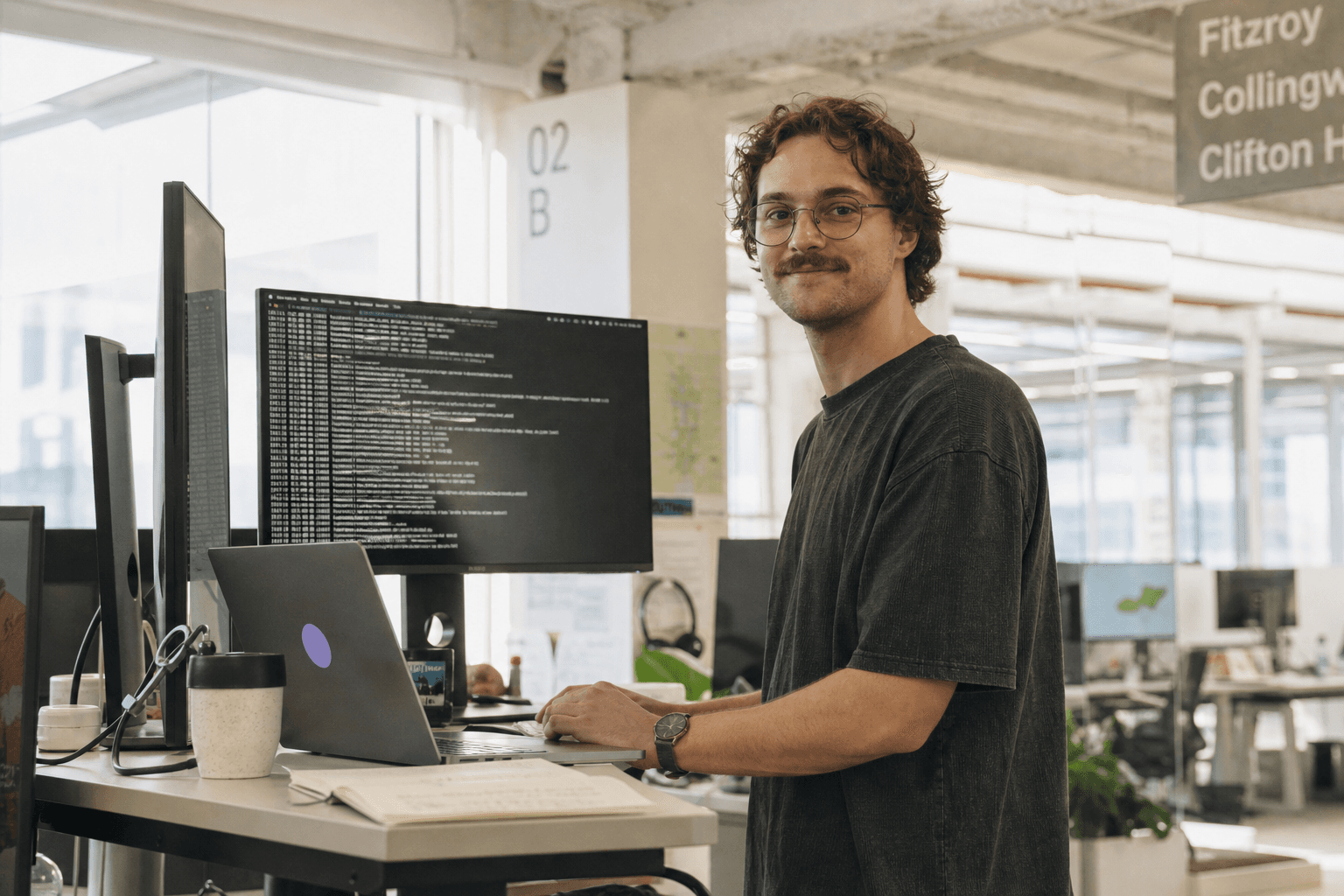 A data engineer standing at a standing desk in a bright, sunlit open-plan office, working at a monitor displaying a terminal window.