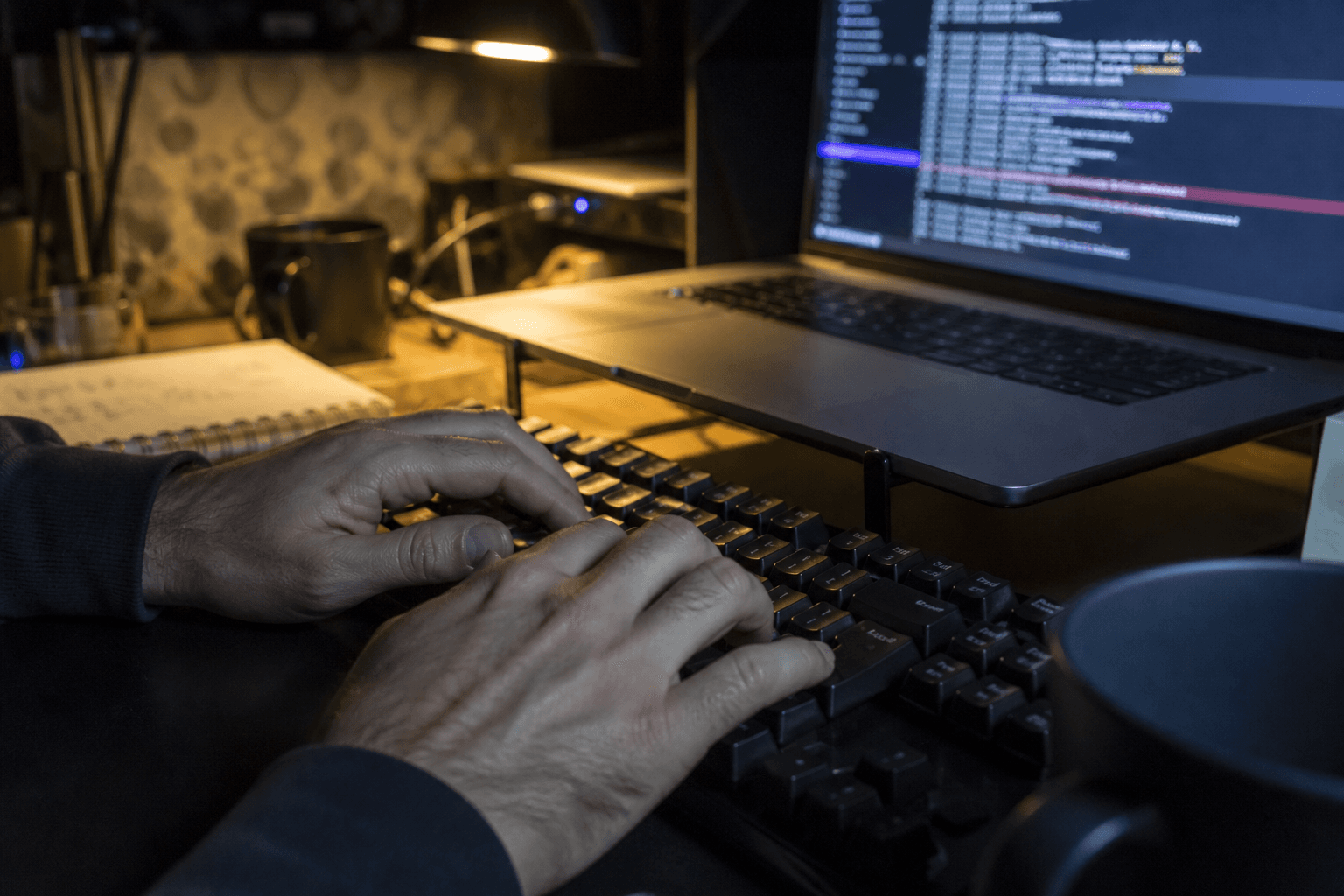 Close-up of hands typing on a keyboard at night, lit by laptop screen glow and a warm desk lamp, with log output visible on the screen.