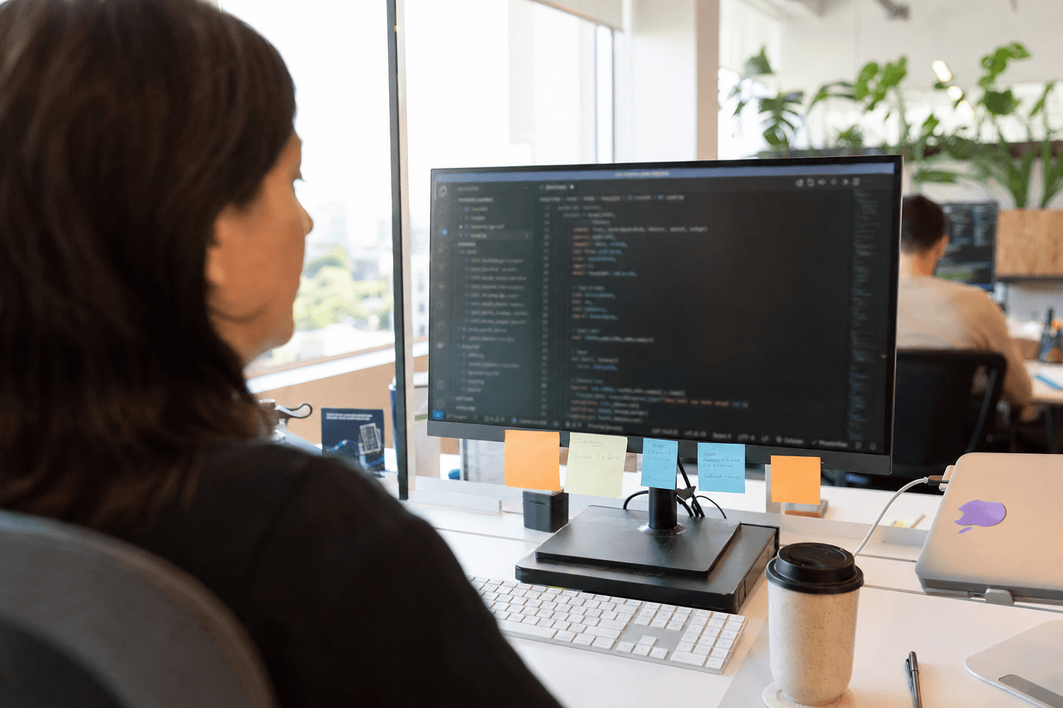 Over-the-shoulder view of a woman at her desk in a bright Australian office, focused on a large monitor showing a code editor, with a sticky-note-covered screen bezel and a coffee cup in the foreground, natural daylight filling the space.