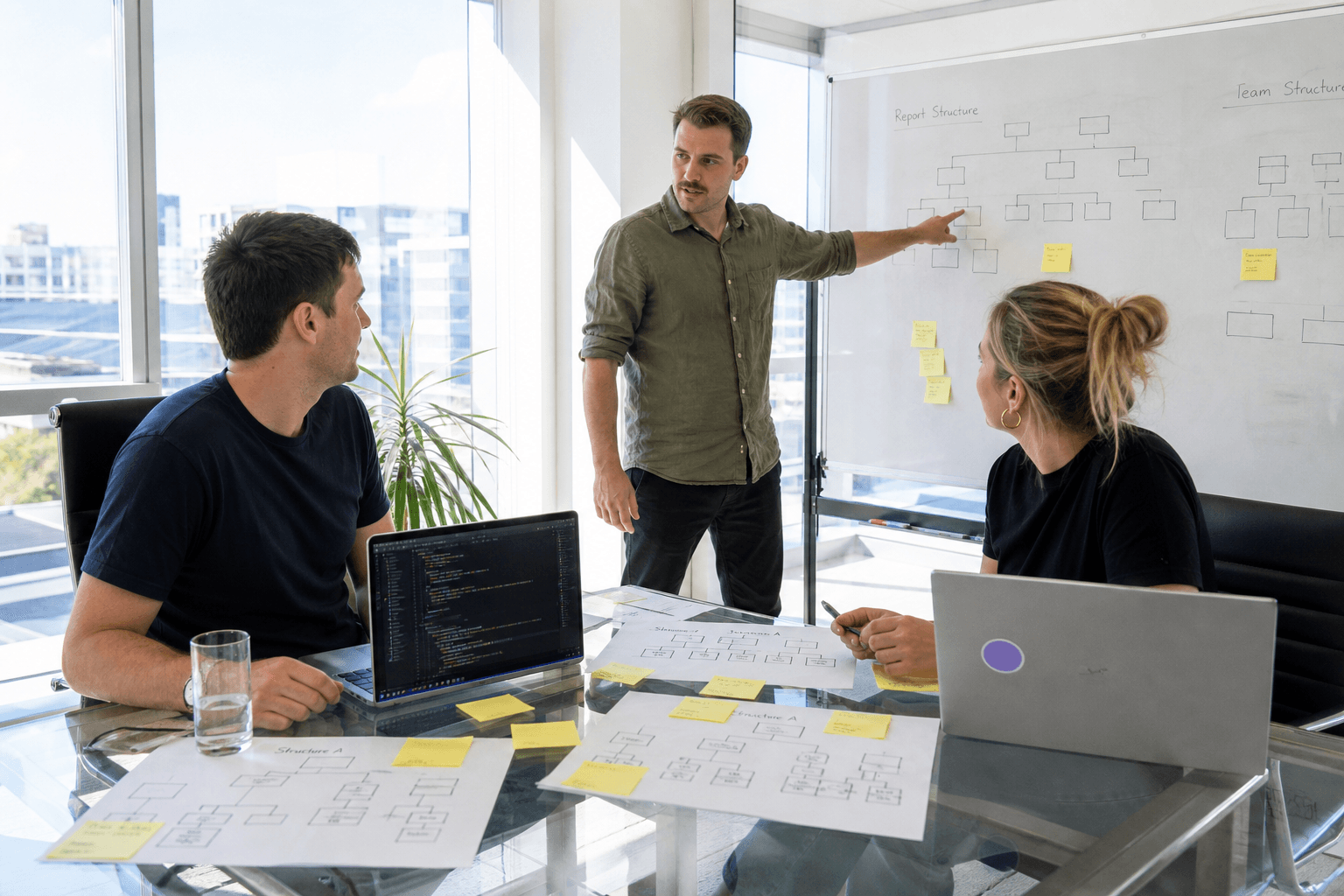 Three colleagues at a glass table in a bright office comparing printed organisational charts and sticky notes, with laptops open and a whiteboard sketch in the background.