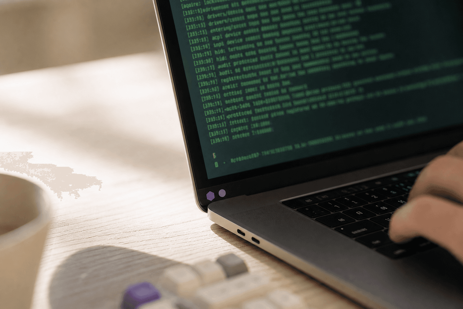 Extreme close-up macro shot of an open laptop screen showing a terminal window, shot at desk level in warm off-white natural office light, with two small violet stickers on the lid and a developer's hand partially visible at the frame edge.