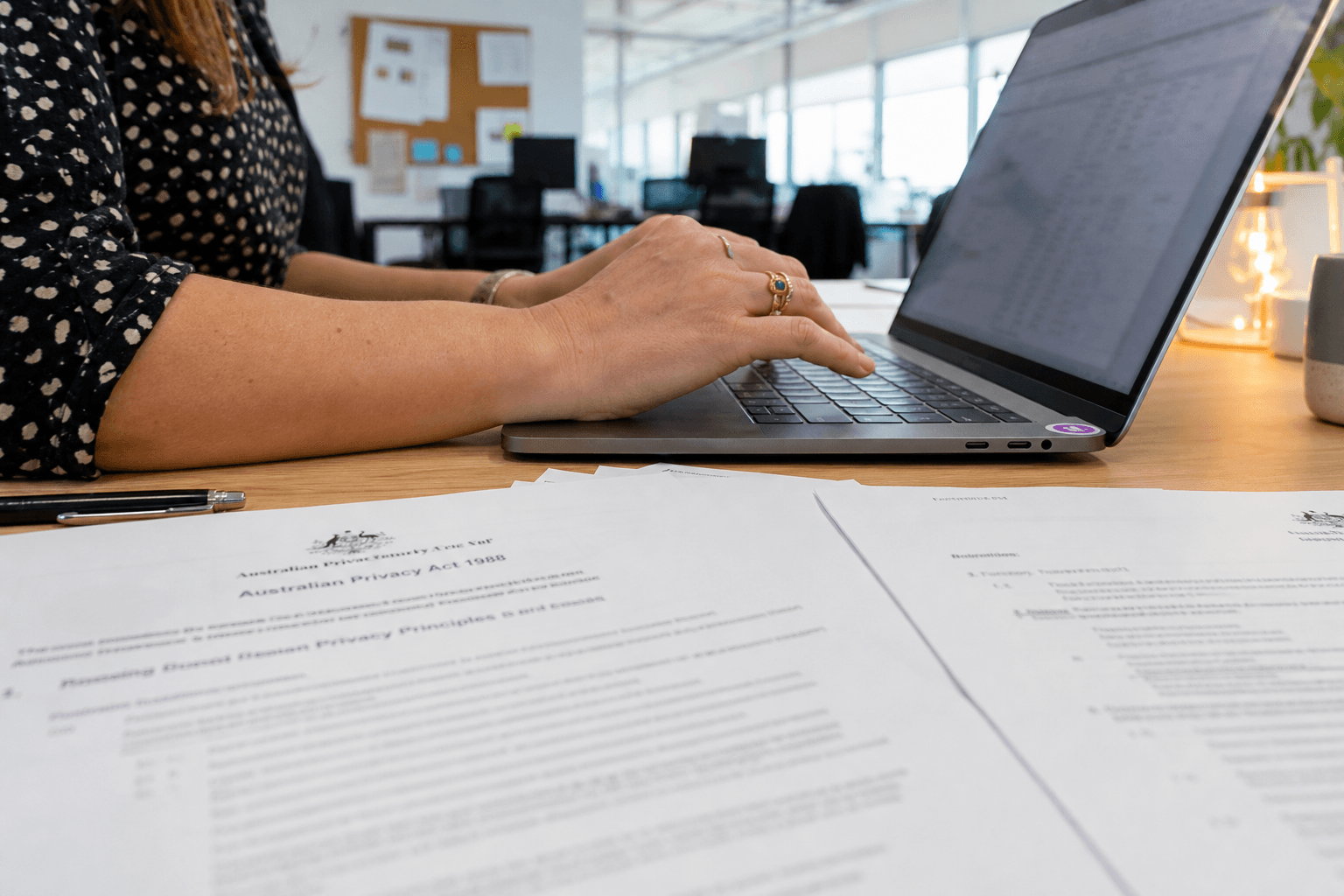 Close-up of a woman's hands on a laptop keyboard with printed compliance documents spread on a light timber desk beside her, photographed in bright natural window light in an Australian open-plan office.