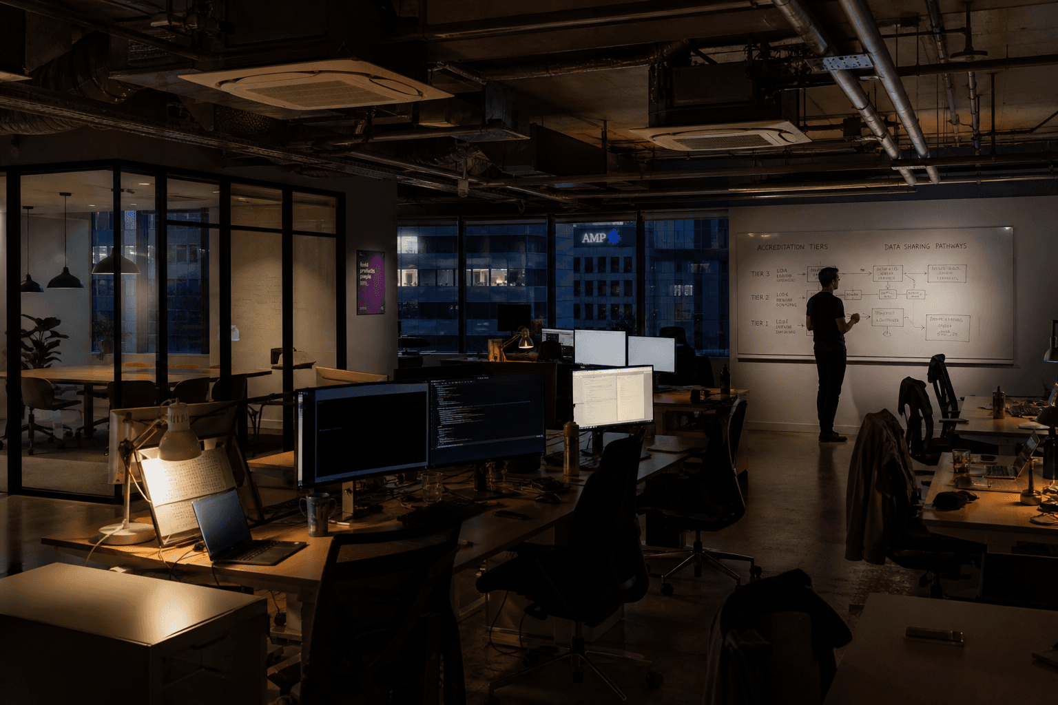 Wide shot of a dimly lit Australian tech office at night, with one engineer standing at a whiteboard reviewing diagrams, the room lit mainly by monitor glow and desk lamps.