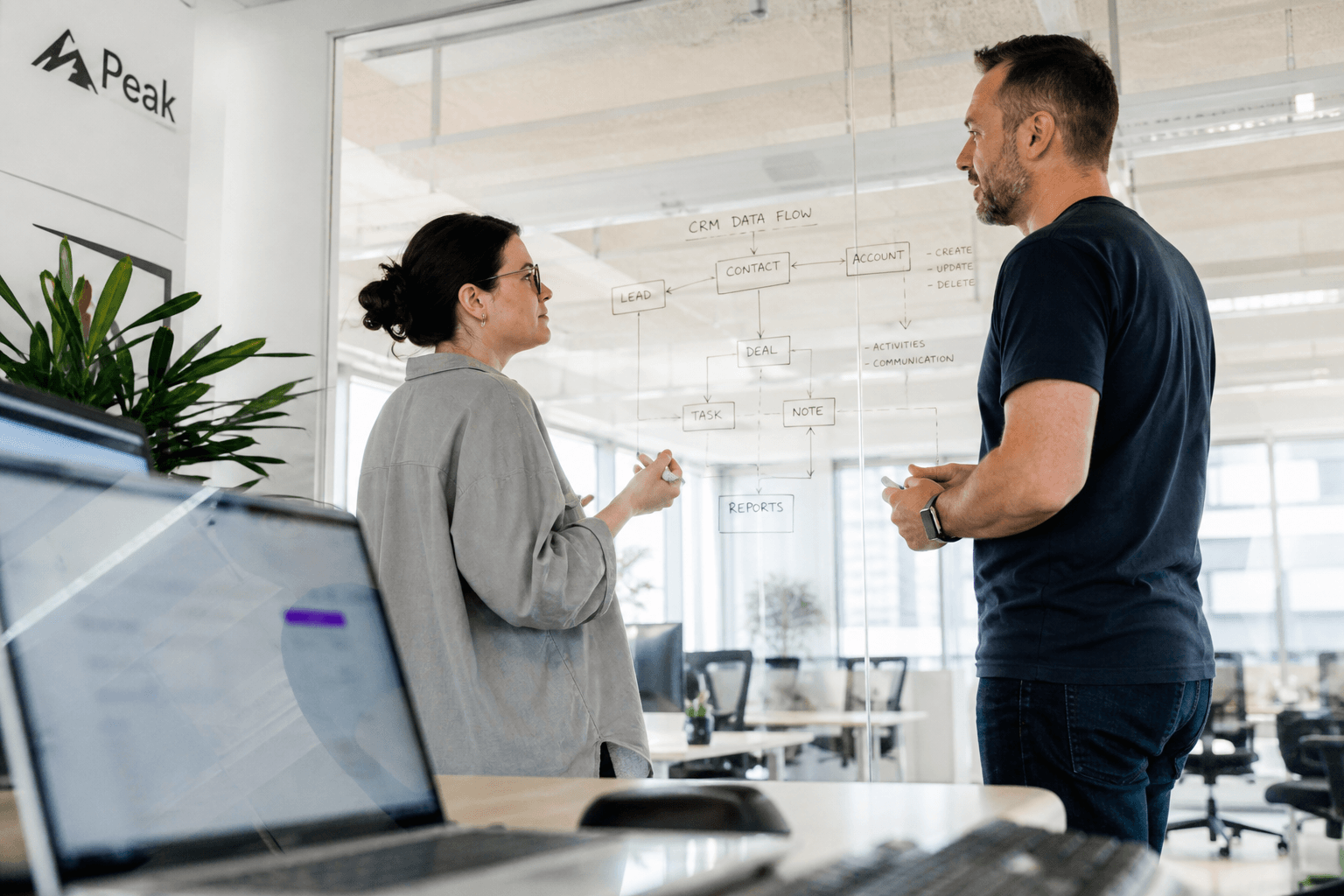 Low-angle view past a laptop toward two engineers discussing a system diagram drawn on a glass wall in a bright, naturally lit Australian office.