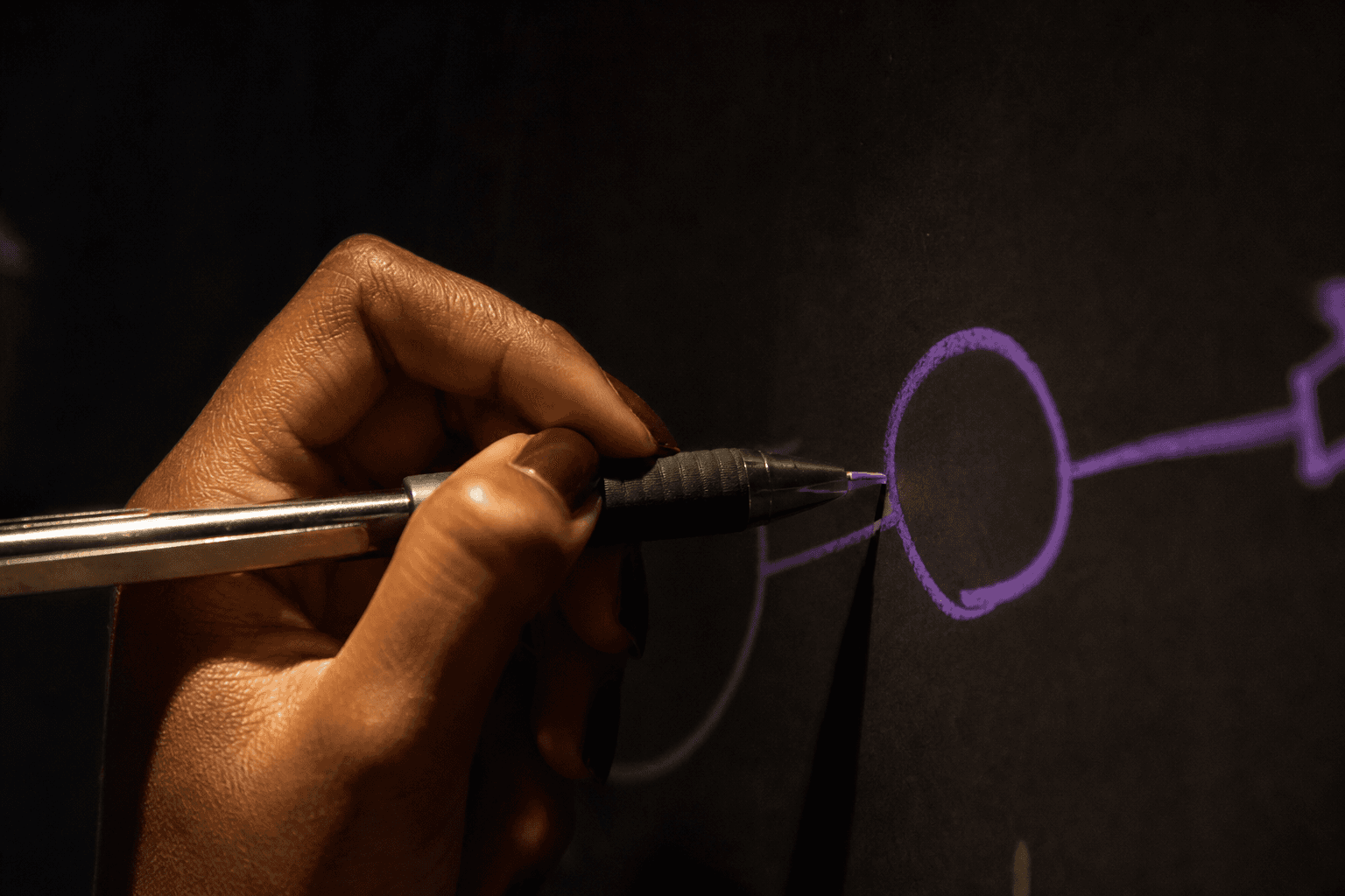Extreme close-up of a dark-skinned hand holding a mechanical pencil, drawing a single circle node on a whiteboard in violet marker. The background is deep shadow, with a narrow strip of warm lamplight illuminating the hand and pencil tip.