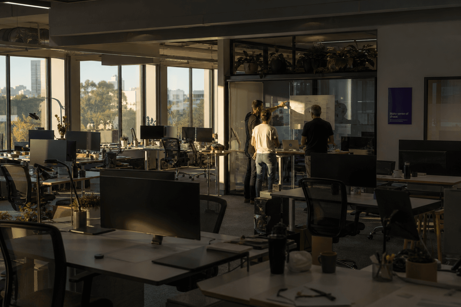 A wide view of an open-plan office at golden hour, showing three engineers gathered around a standing desk looking at a printed roadmap taped to a glass partition, with rows of empty desks in the background.
