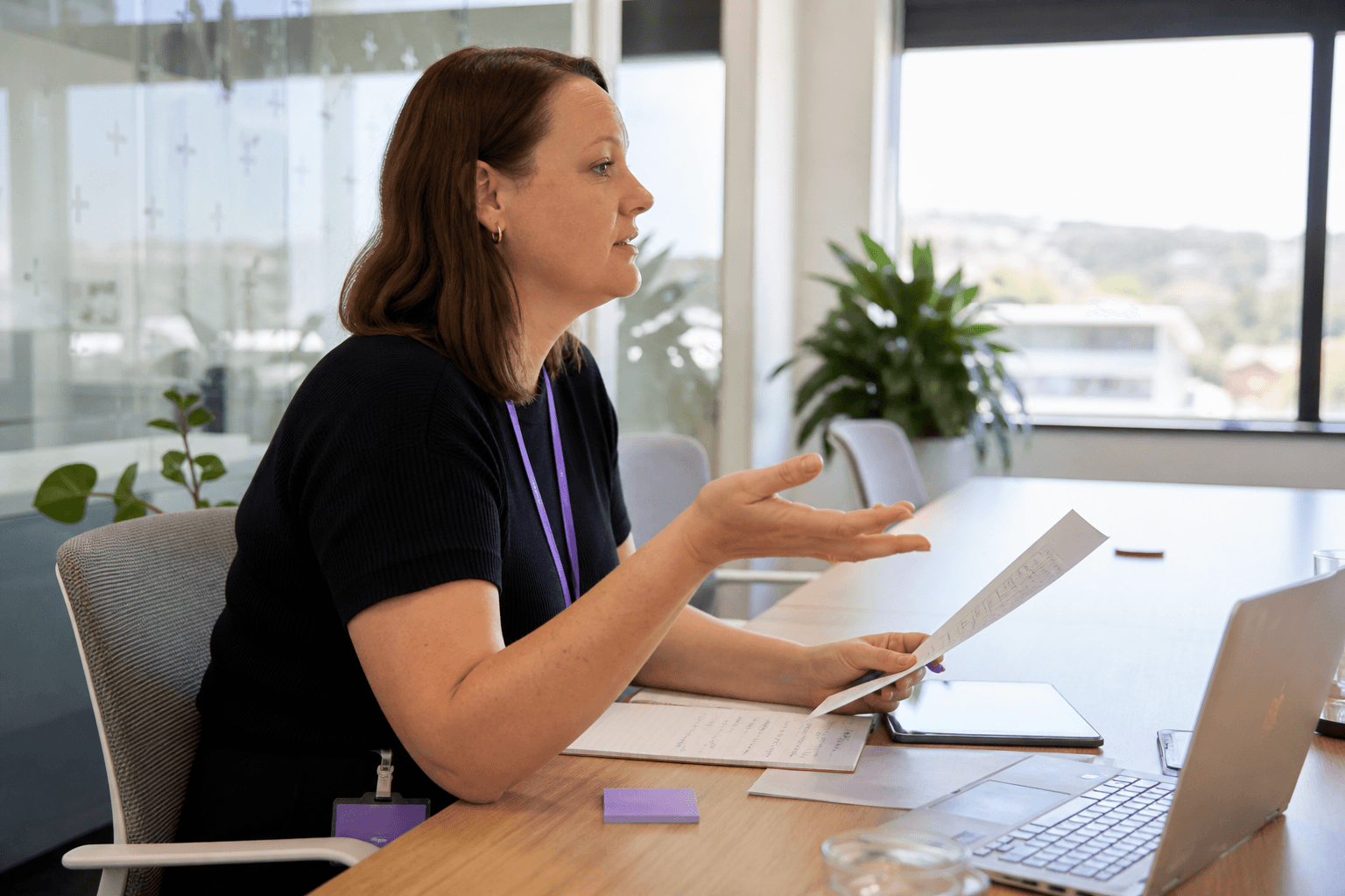 A professional is caught mid-conversation in profile at a bright meeting table, reviewing printed documents and a laptop in a sunlit office.