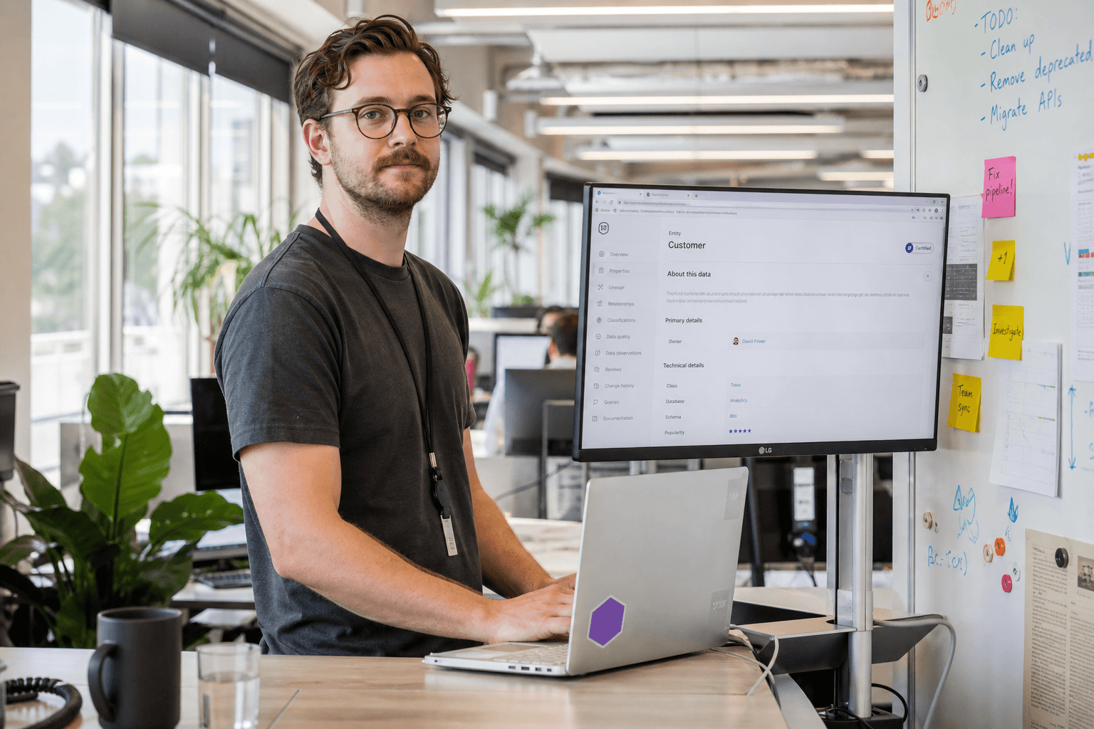 A data engineer standing at their desk in a bright, sunlit open-plan office, with a monitor showing a data catalogue page and sticky notes on a whiteboard nearby.