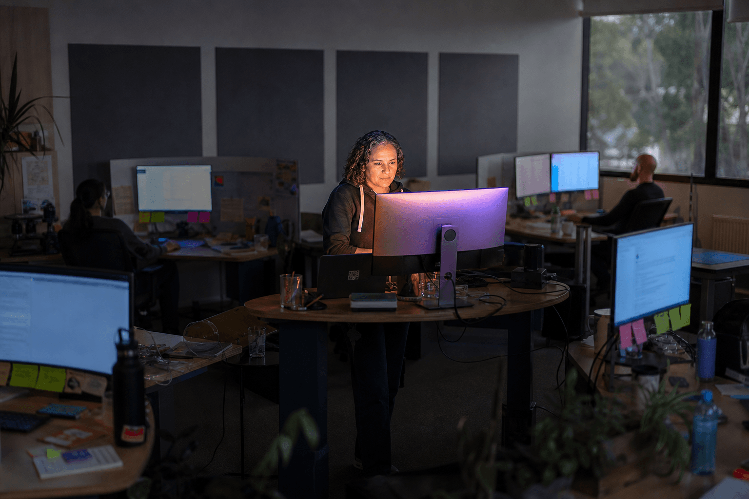 A senior engineer sits at a central standing desk in focus, surrounded by three separate disconnected workstations arranged in an orbital pattern at the edges of the frame, each with isolated cold monitor glow, in a mid-lit Australian office.