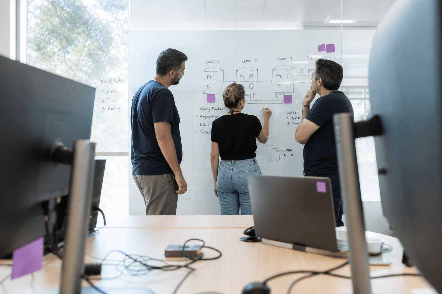 Engineers sketch a distributed database diagram on a glass whiteboard, seen through the gap between two monitors on a bright, daylit desk.