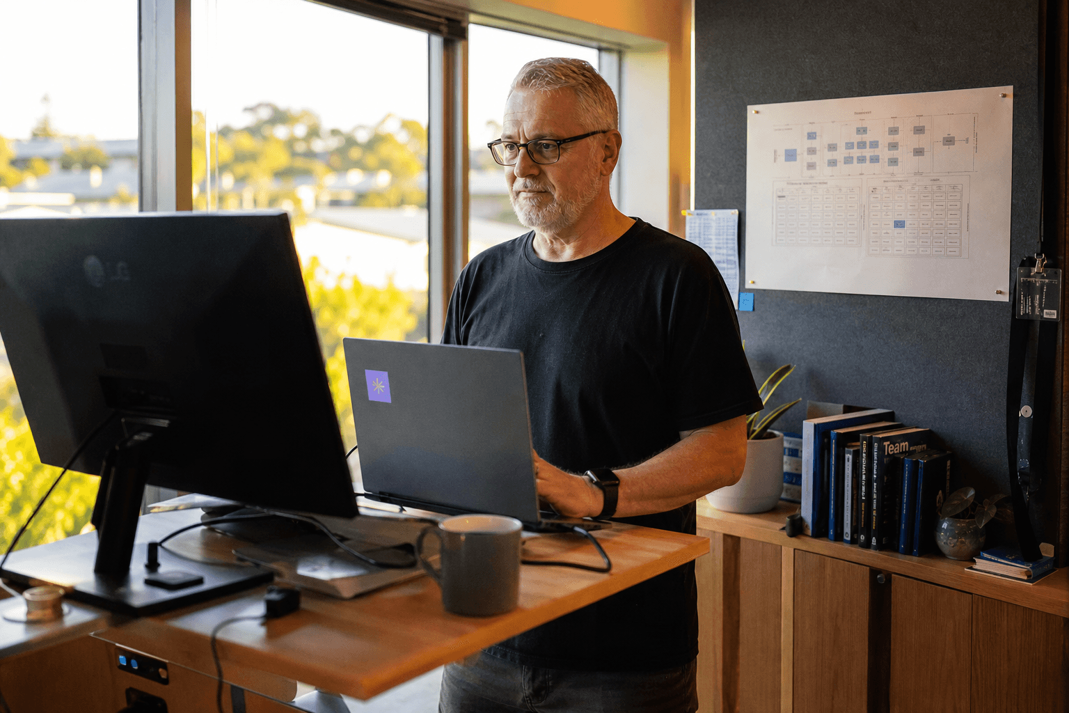 A senior engineer stands at a standing desk workstation with a printed architecture diagram on the wall behind them, lit by warm golden-hour window light.