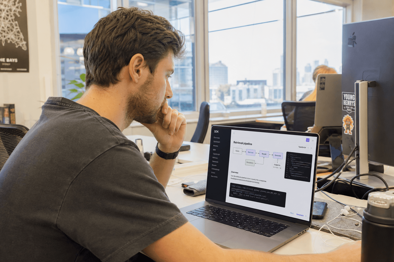 Side profile of a person sitting at a desk in a bright, sunlit office, looking at a laptop screen showing documentation and a diagram.