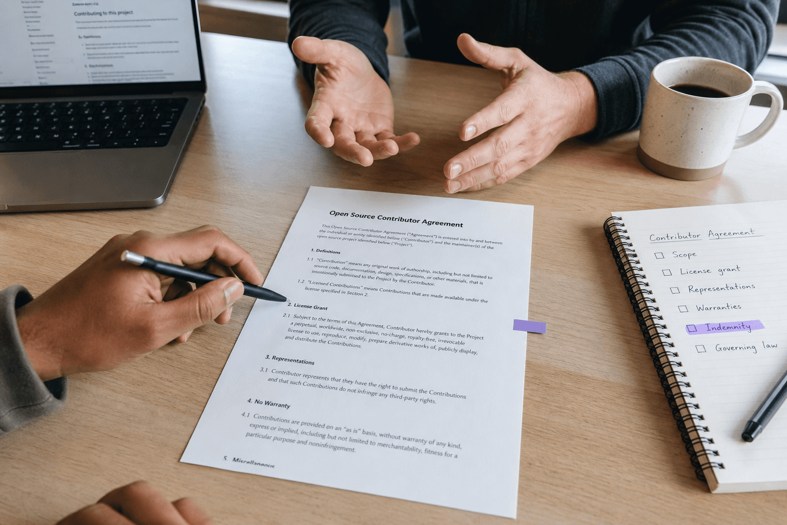 Overhead view of a printed contract document on a light timber desk surrounded by a pen poised above the page, open hands across the table, an open laptop, a coffee cup and a notepad with a handwritten checklist, lit by cool window daylight.