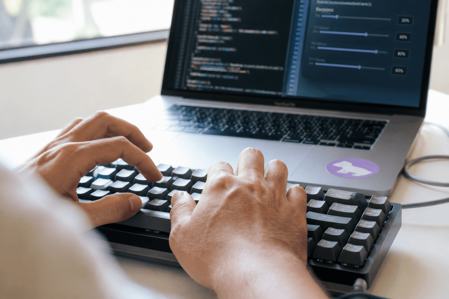 Close-up of hands typing on a mechanical keyboard in bright daylight, with a laptop screen showing code and a toggle interface softly out of focus behind.