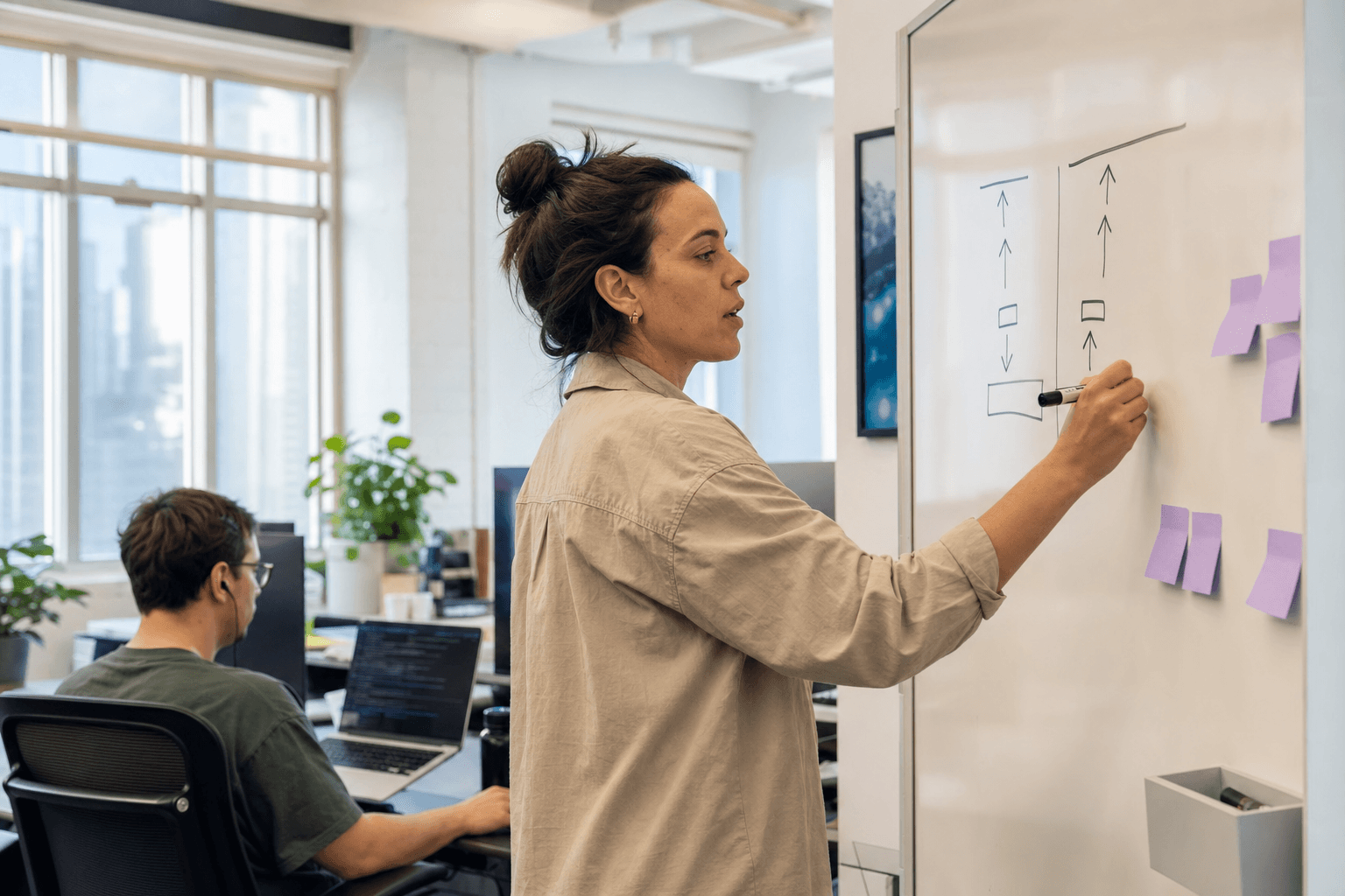 A female ML engineer writes a model size comparison diagram on a whiteboard in a bright Australian tech office, viewed from the side in a candid unposed moment, with a colleague visible at a laptop in the background.