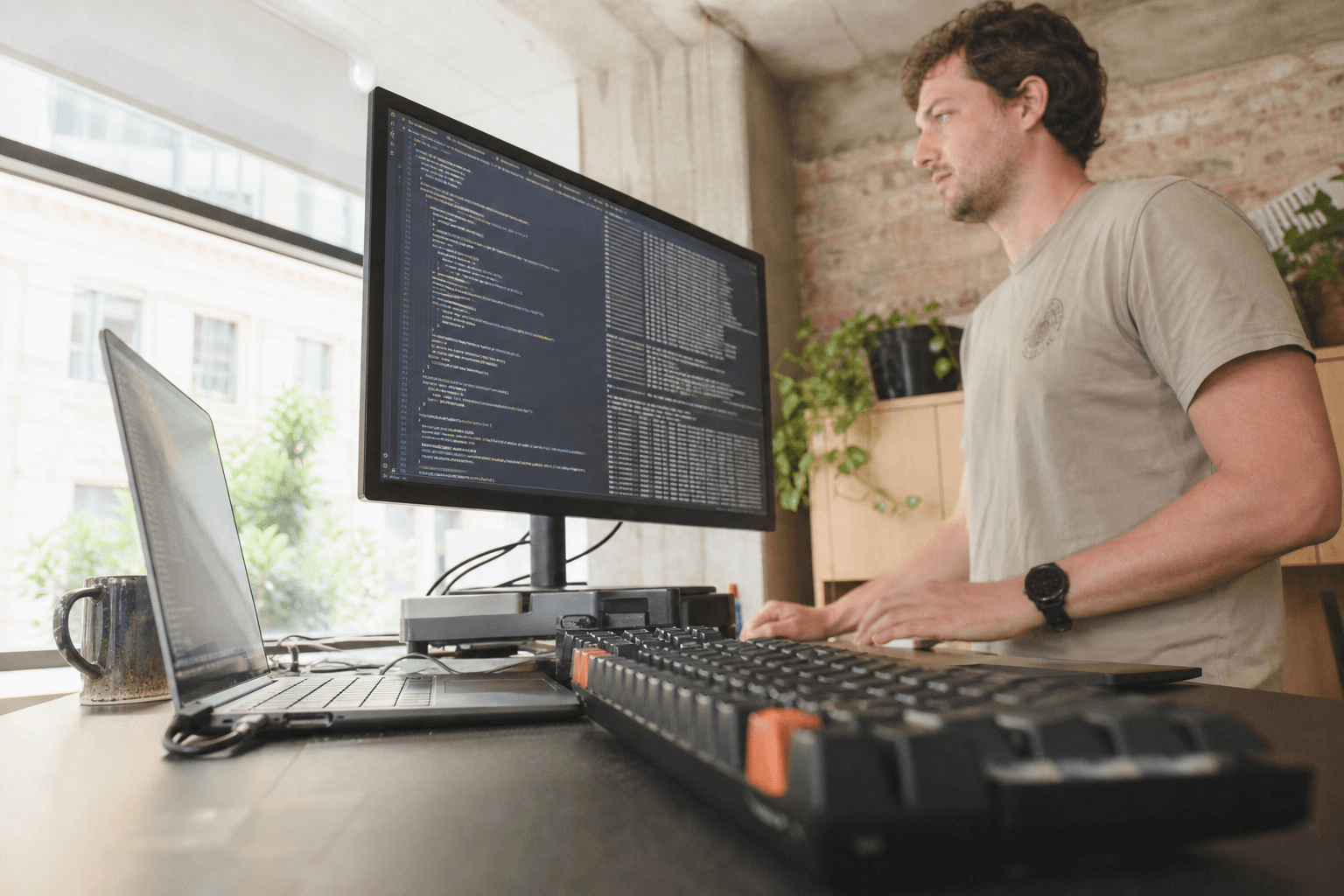Low-angle view from near a desk surface looking up past a keyboard and laptop toward a developer standing at a monitor displaying code columns, in a bright daylit Australian office interior.