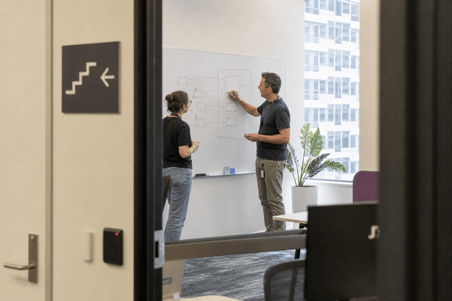 Two colleagues in an Australian office viewed through a doorway, standing at a whiteboard with a simple hand-drawn diagram of boxes and arrows, lit by bright daylight.