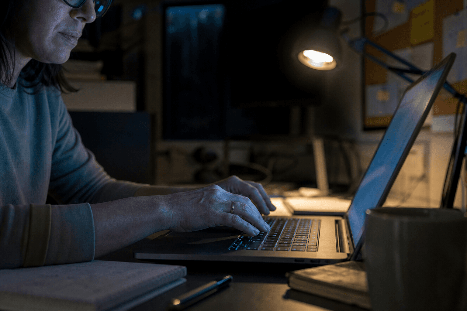 Close-up of a woman's hands typing on a laptop keyboard in a dimly lit office, bathed in cool screen glow with warm task lamp light from behind, shallow depth of field with soft-focus desk and corkboard in the background.