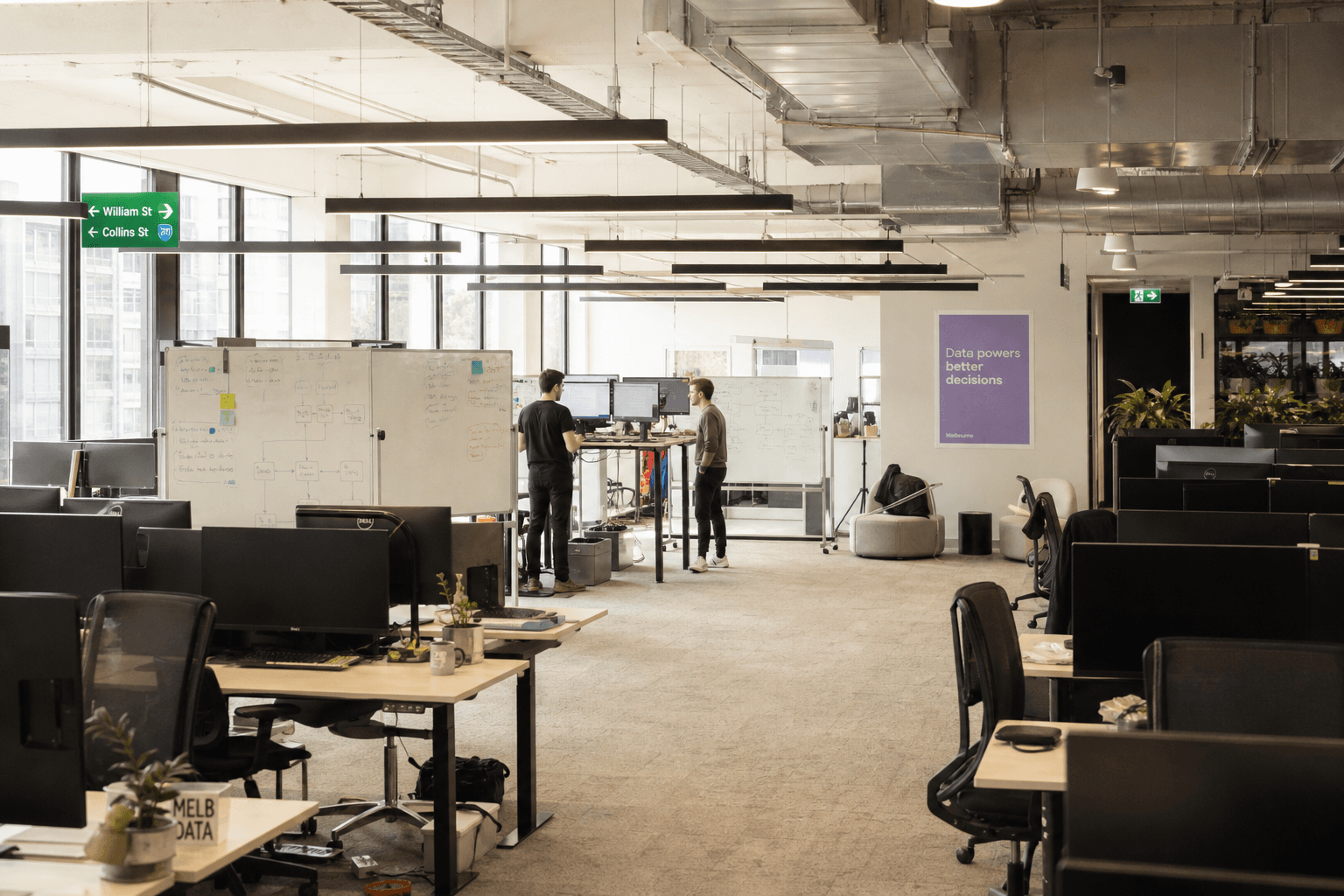 A wide view of a bright, open-plan office with two data engineers working at a standing desk near whiteboards covered in system diagrams, daylight streaming through large windows.