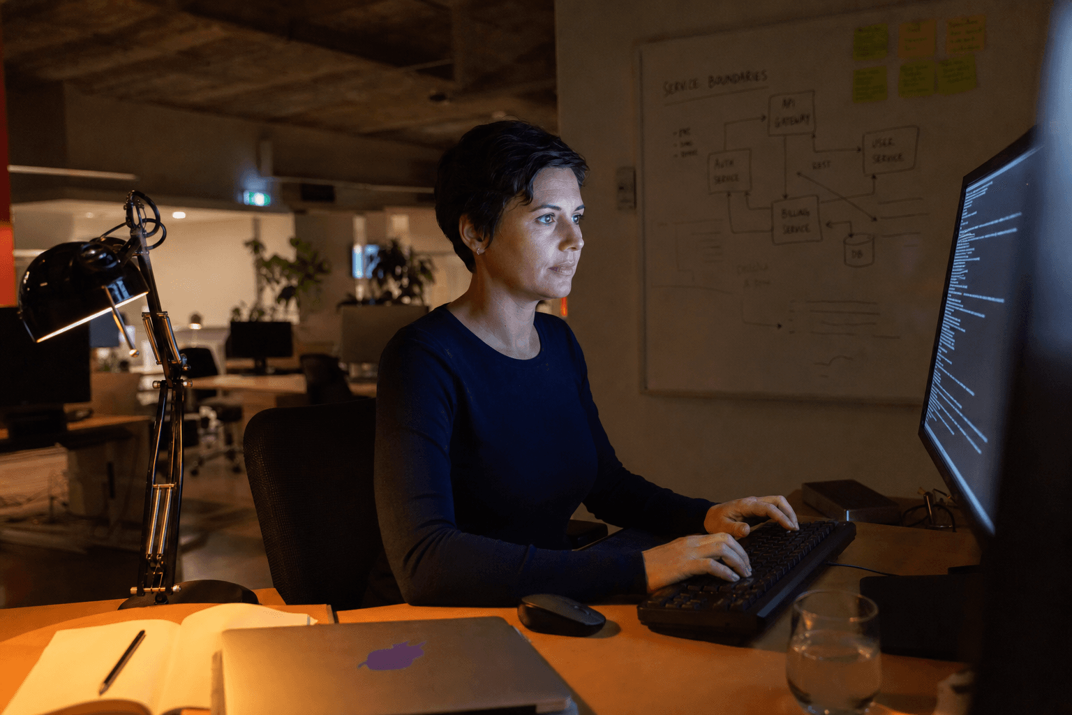 A software engineer sits at a desk in a dimly lit Australian tech office at night, her face lit by warm task lamp glow and cool monitor light, with a whiteboard showing service-boundary sketches softly blurred in the background.