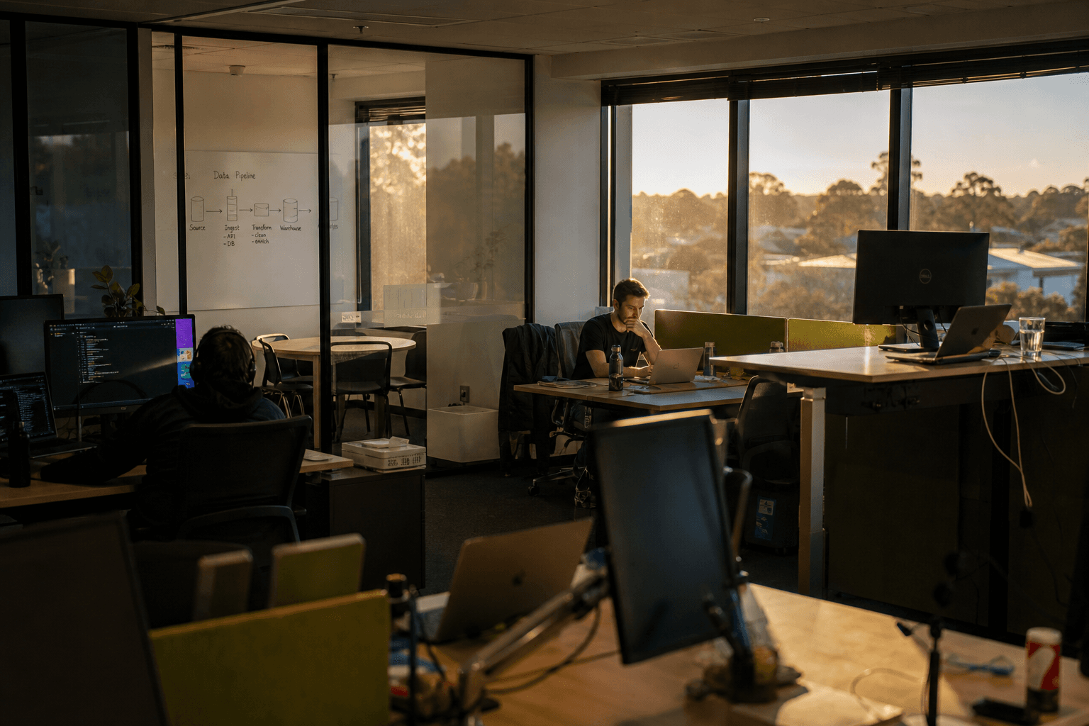 Wide view of an open-plan Australian office with a small figure working at a laptop near sunlit windows, desks and a whiteboard with a simple diagram visible in the background.