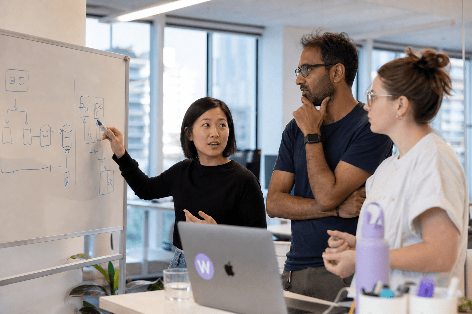 Three colleagues in bright daylight standing around a whiteboard with a simple hand-drawn diagram, discussing and pointing at the sketch in an open office.